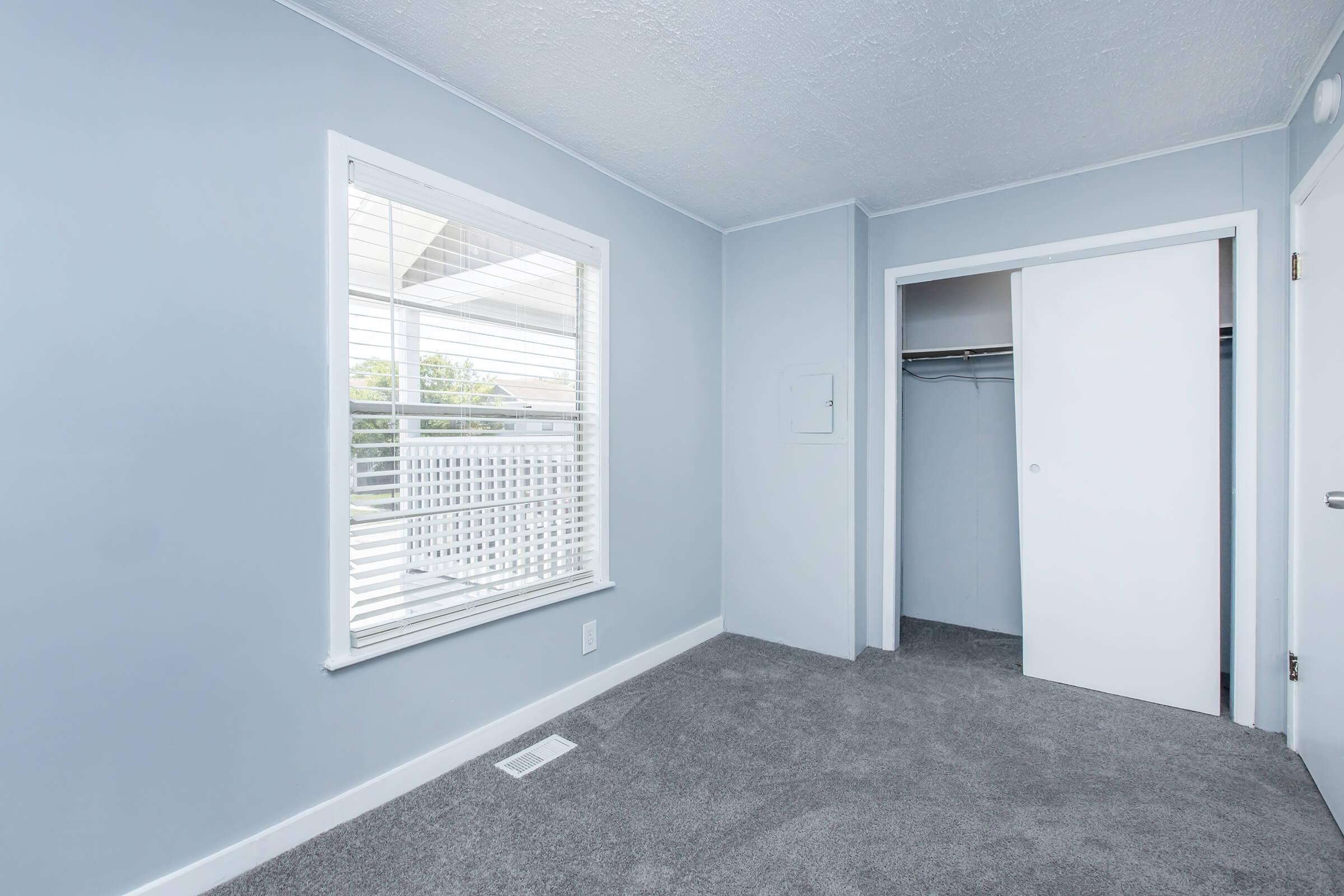 A light blue room with a window featuring white blinds. The room has gray carpet and a closet with sliding white doors on the right. Sunlight enters through the window, creating a bright and airy atmosphere.