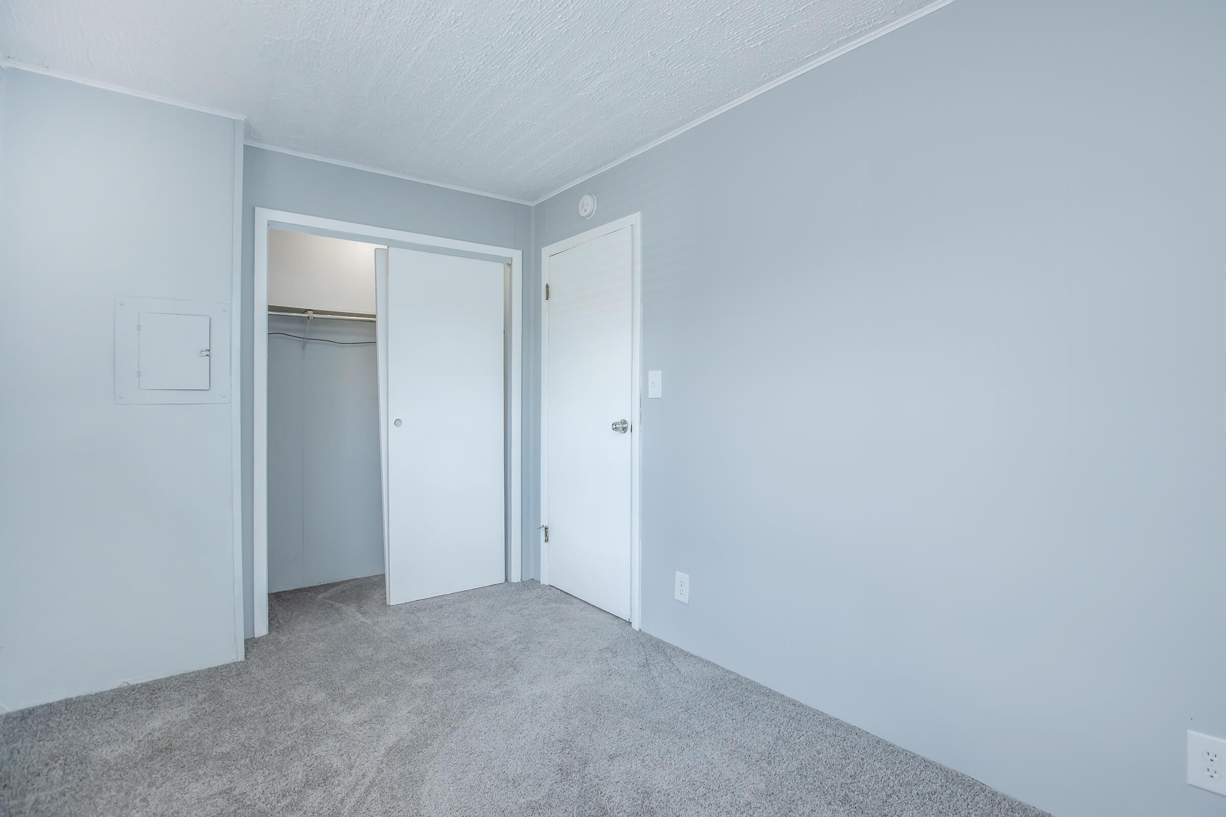 A spacious room with grey walls and carpeted flooring. There are two doors: one leading to a closet and another to a bathroom or hallway. The room is well-lit, featuring a textured ceiling, and the overall atmosphere is quiet and minimalistic.