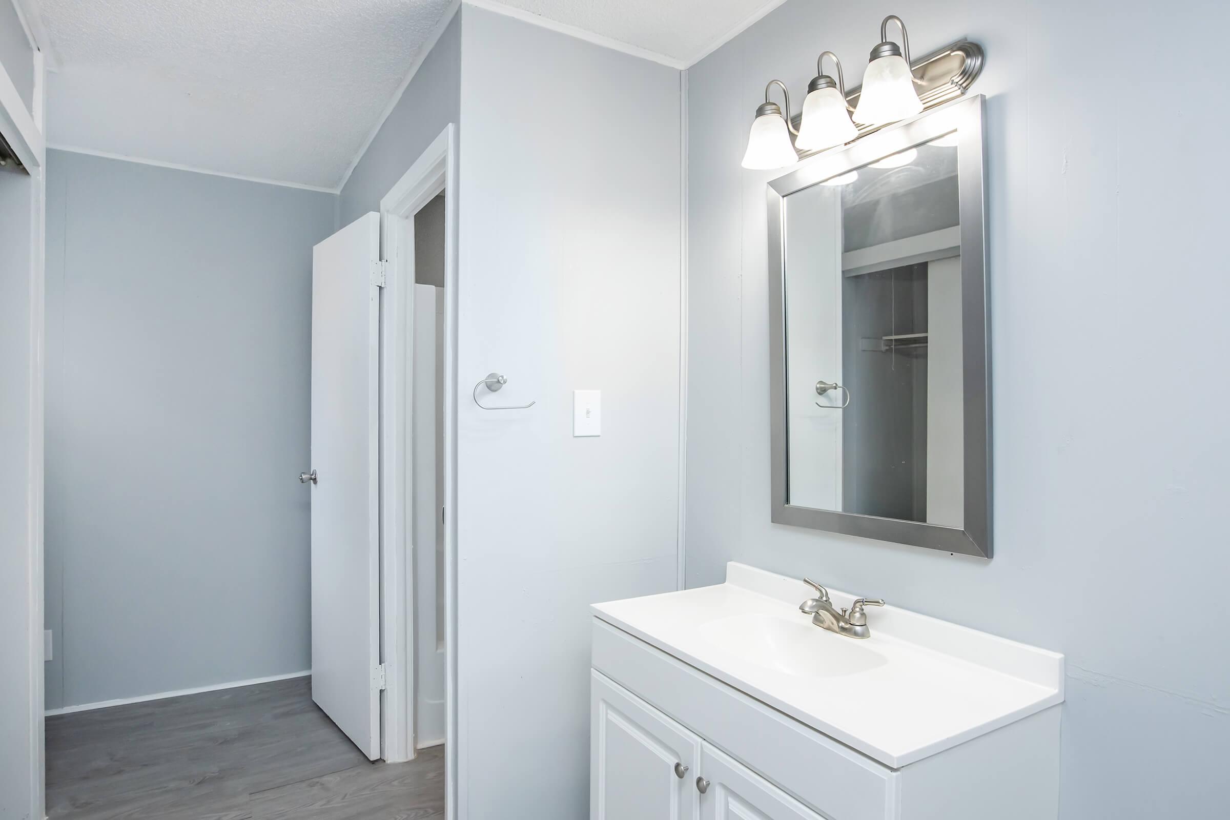 A clean and modern bathroom featuring a white vanity with a sink, a large mirror above it, and stylish light fixtures. Light gray walls create a soft ambiance, and a door on the right leads to another room. Light wood flooring adds warmth to the space.