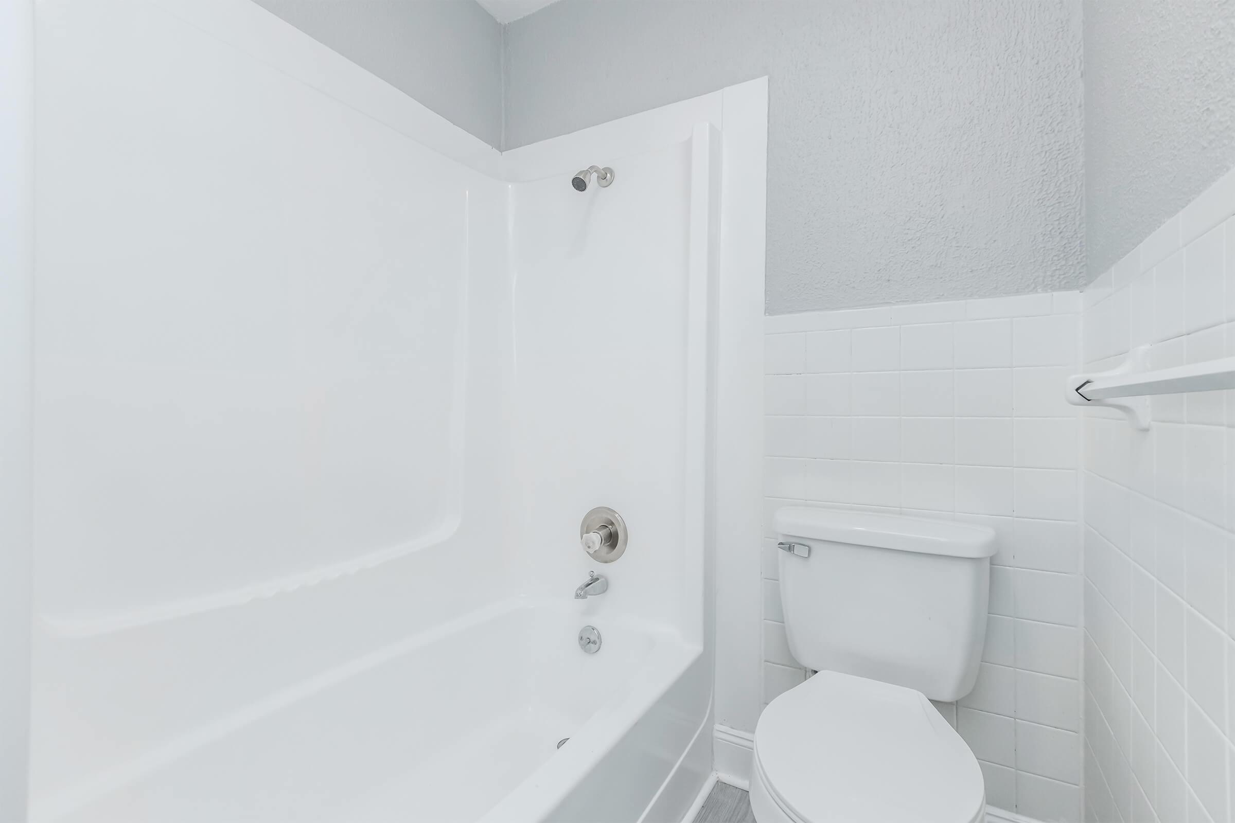 A clean and simple bathroom featuring a white bathtub with a showerhead, a white toilet, and light gray walls. The floor is tiled, and the space is well-lit, giving it a fresh and modern appearance.