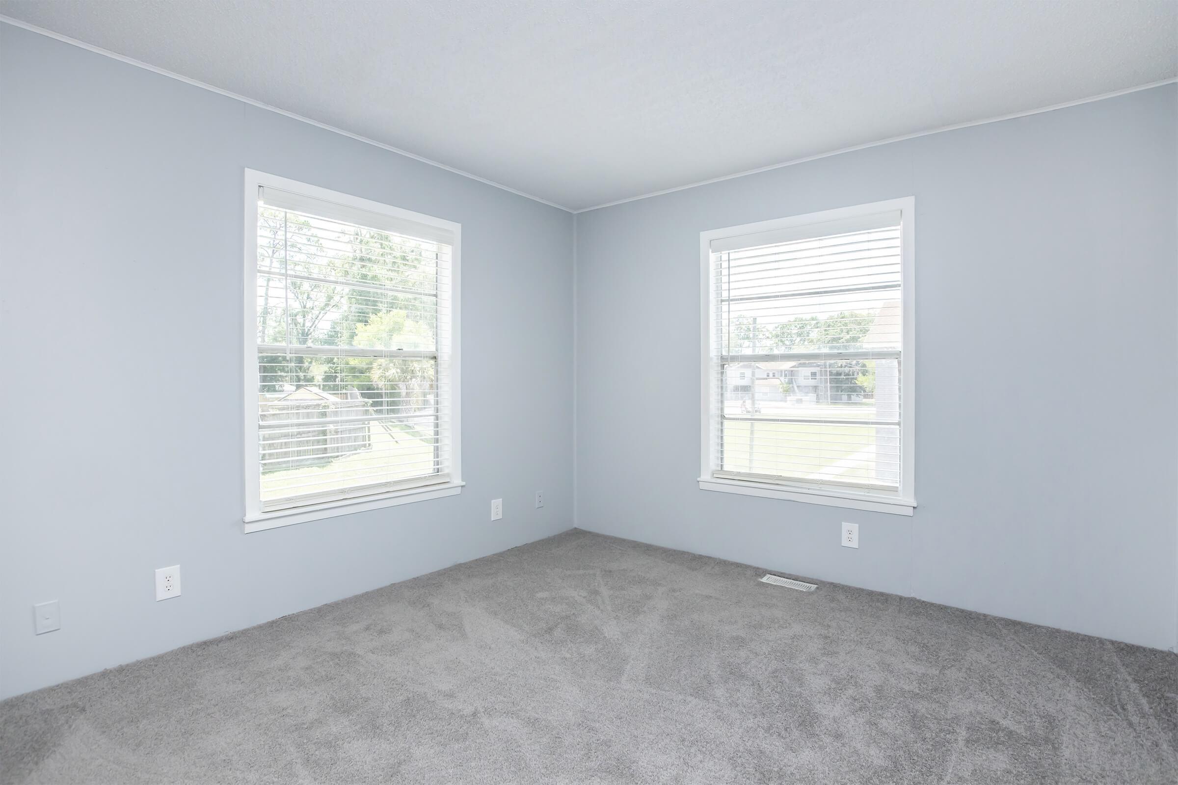 Empty room with light gray walls and two windows on adjacent walls, featuring white blinds. The floor is covered in a soft, light gray carpet. Natural light enters through the windows, illuminating the space, which has a minimalistic and clean appearance. There are no furnishings or decorations present.