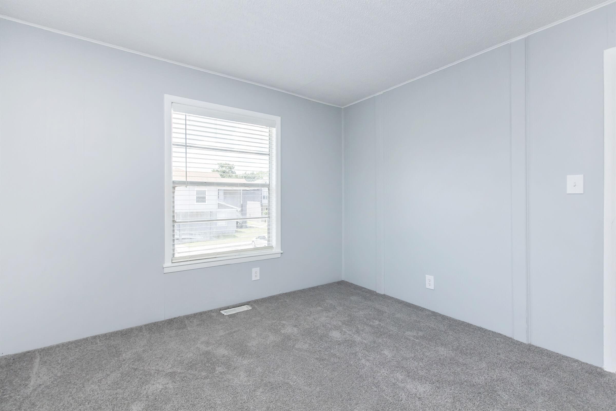 A bare room with light gray walls and soft gray carpet. There is a window with white blinds allowing natural light, and the room has a clean, minimalist look. A single electrical outlet is visible on one wall. The space appears empty and ready for furnishing.