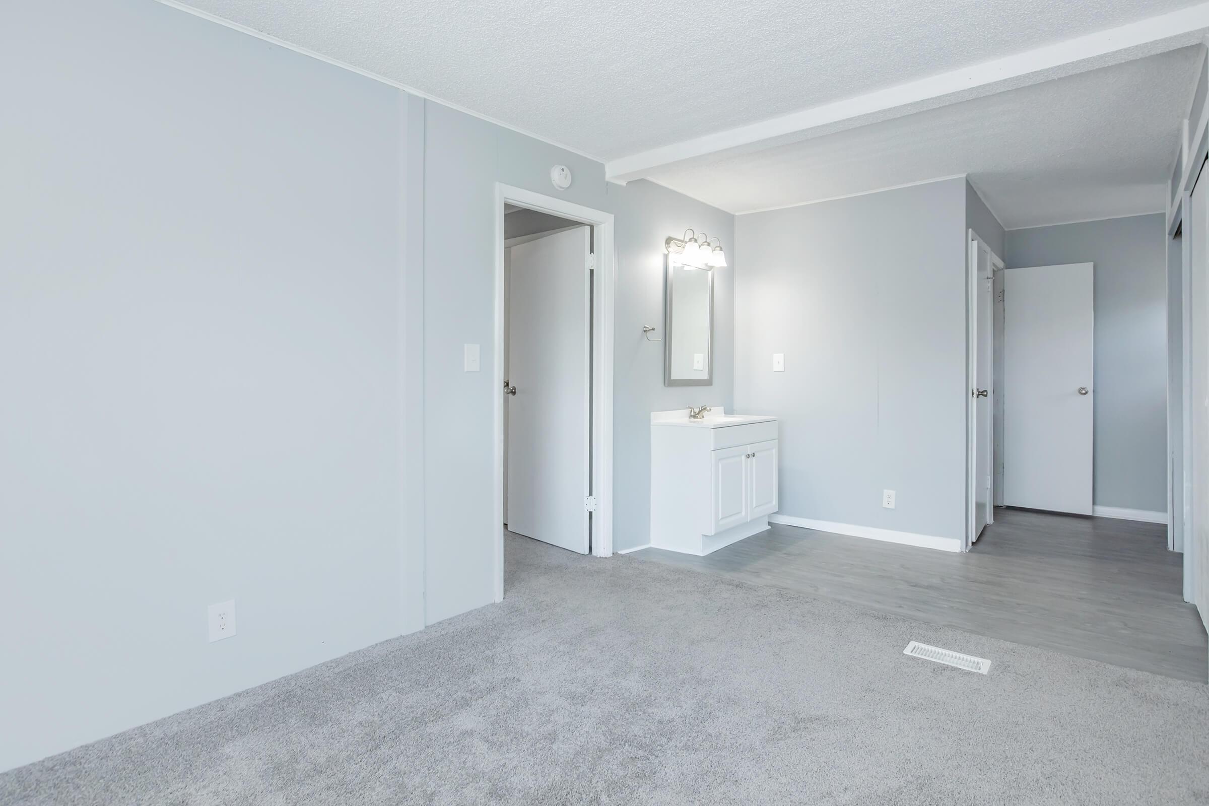 A well-lit, empty room featuring light gray walls and carpet. There is a doorway leading to another room, a small vanity with a mirror on one wall, and two closed doors on the right side, suggesting a simple, modern design.