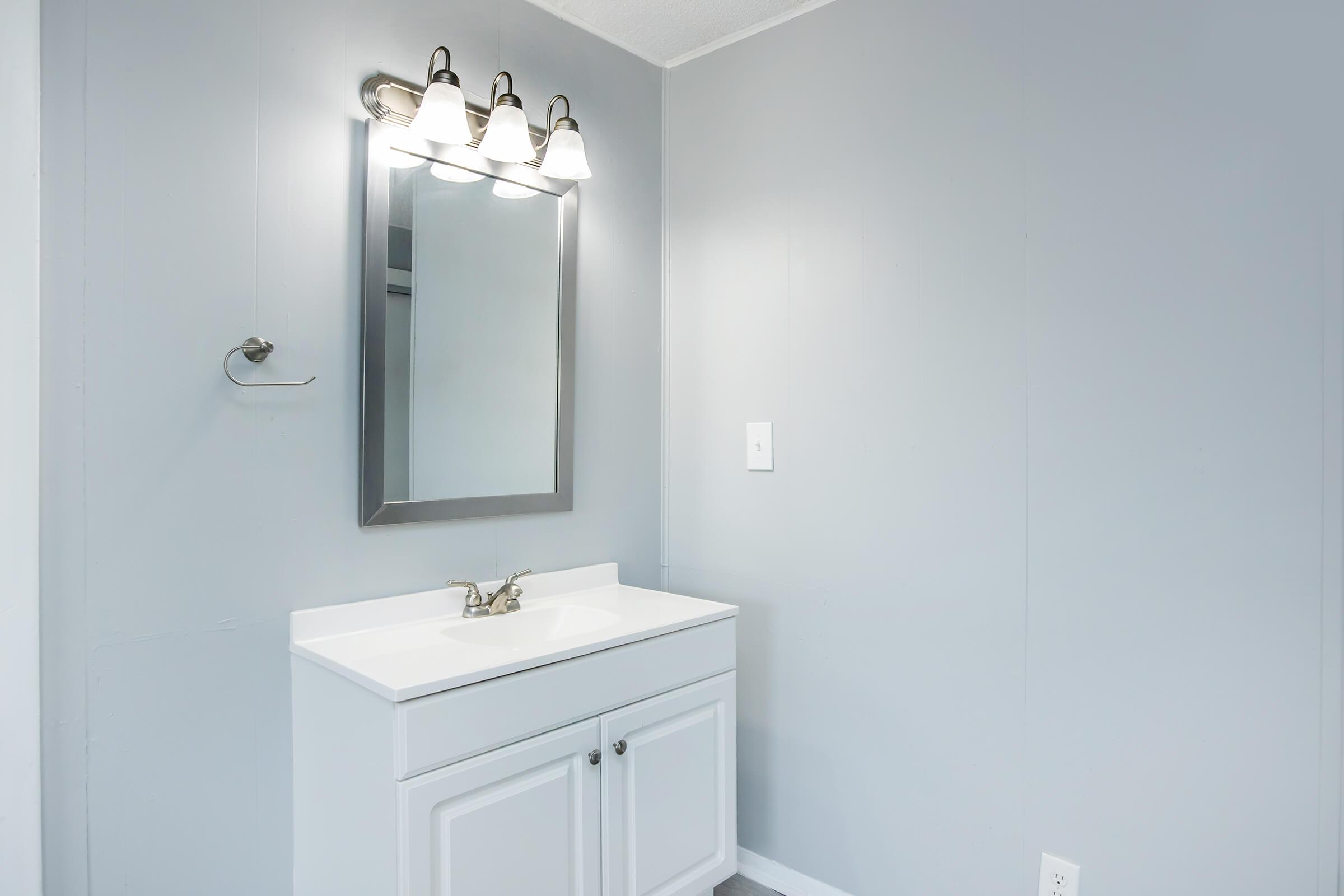A modern bathroom vanity with a white countertop and cabinet, featuring a rectangular mirror above. There are three light fixtures mounted on the wall above the mirror, and the walls are painted in a light gray color, creating a clean and minimalist design.