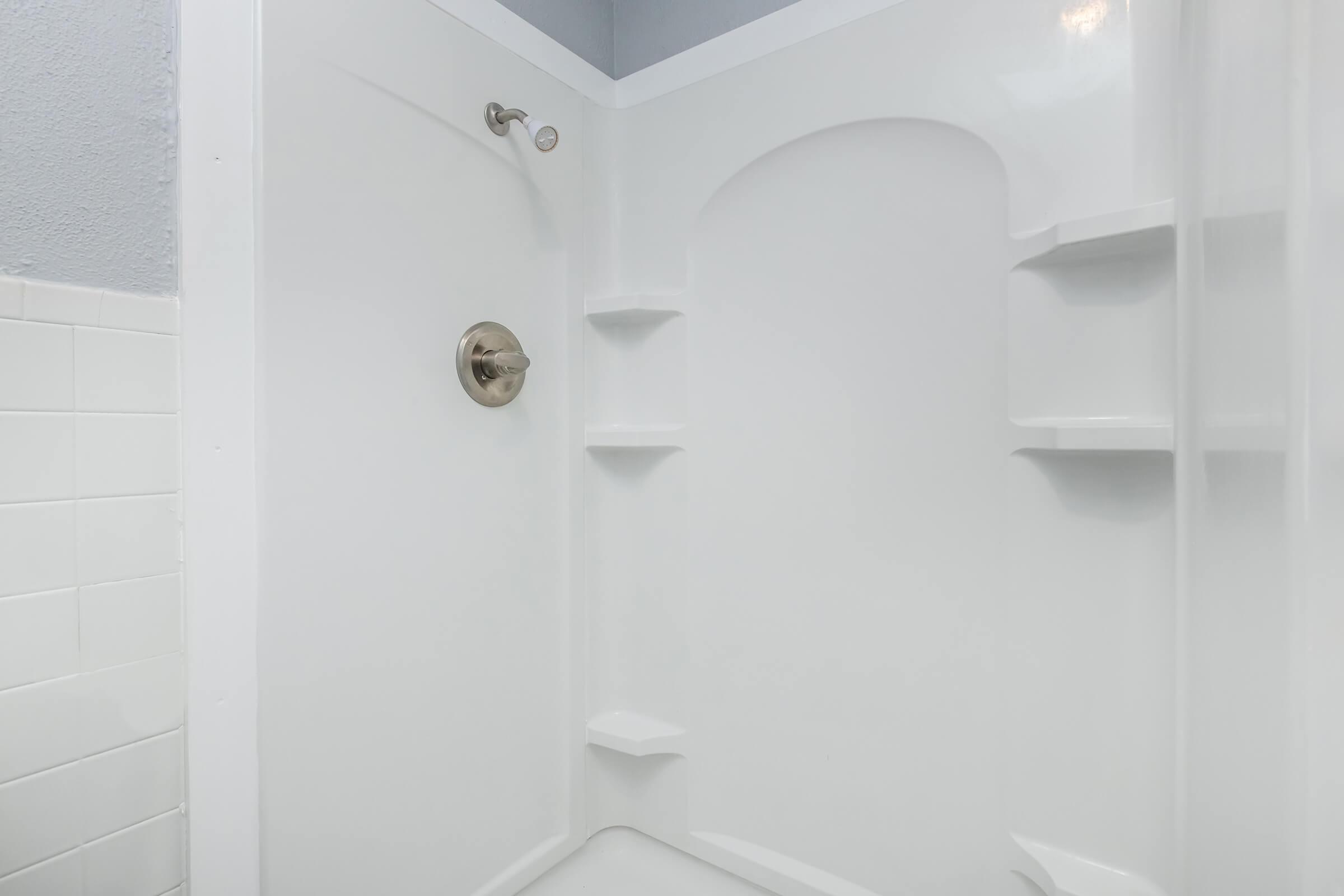 A clean, modern shower stall featuring white walls, a smooth, curved design, and built-in shelves. The fixture includes a chrome faucet and showerhead, with a light blue accent wall above. The overall look is minimalist and bright, ideal for a contemporary bathroom setting.