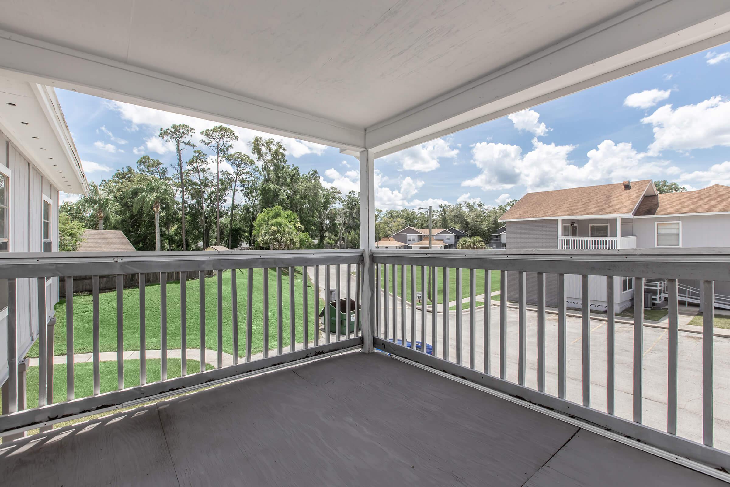 View from a covered balcony overlooking a green lawn and nearby houses on a sunny day, with scattered clouds in a blue sky. The balcony features a wooden railing and is shaded, providing a cozy outdoor space.