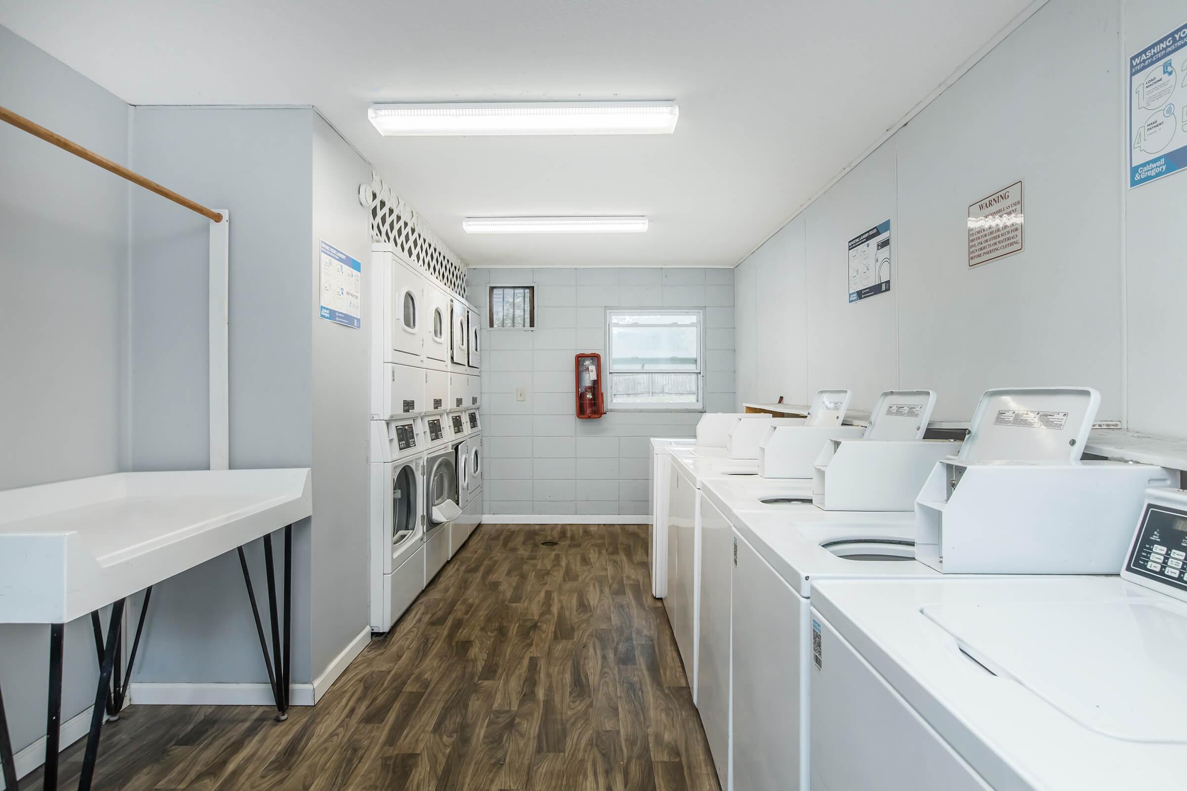 A clean and spacious laundry room featuring multiple washing machines and dryers arranged along the walls. There's a folding table on the left side and a window providing natural light. The walls are light-colored, and safety equipment is visible on the wall.