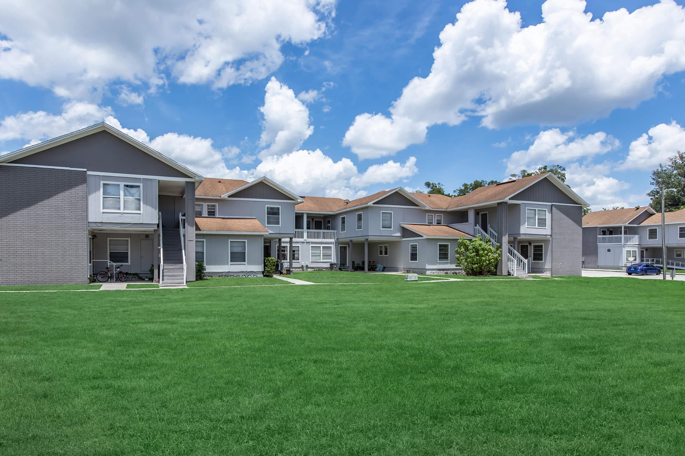 A view of a residential complex featuring multiple gray apartment buildings with brown roofs, set on a well-maintained green lawn under a bright blue sky with fluffy white clouds. The area appears serene and inviting, with several walkways leading between the buildings.