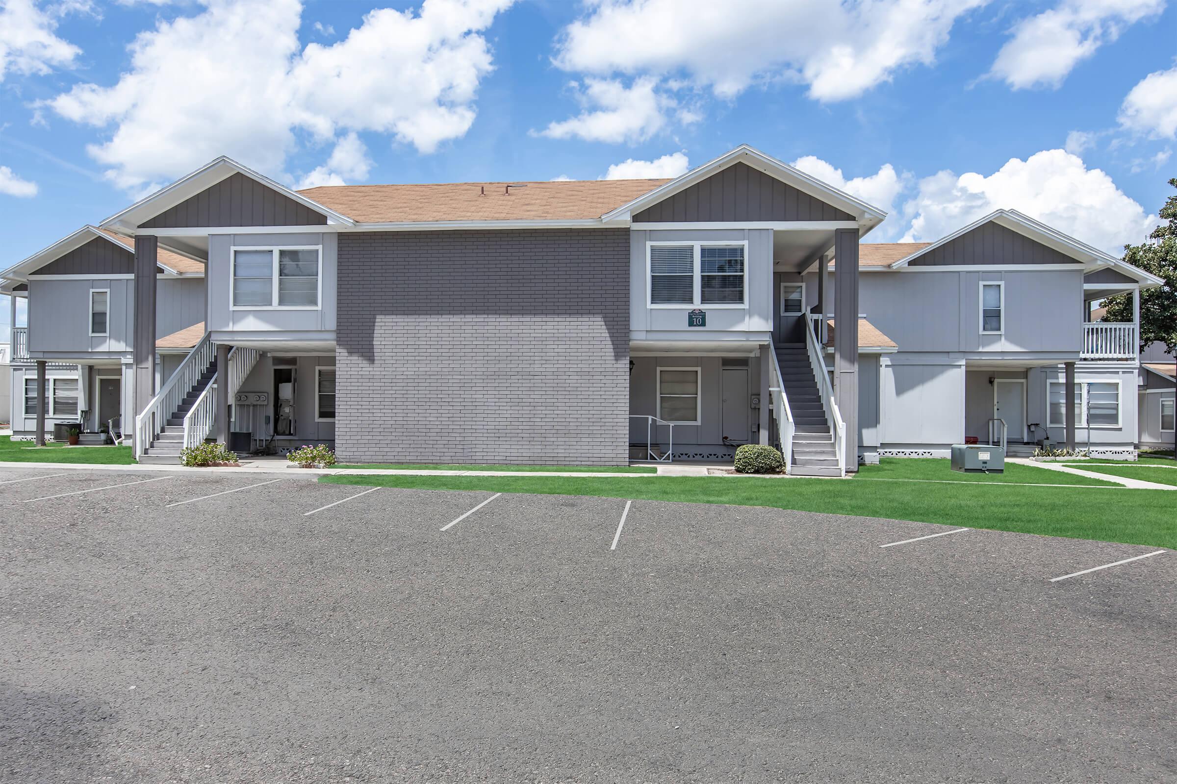 A multi-unit apartment complex with a modern design featuring two stories. The exterior has a mix of gray and light colors, with multiple entries and balconies. In the foreground, there is a paved parking area with several empty parking spaces, and green grass landscaping surrounding the building. Bright blue sky with fluffy clouds above.