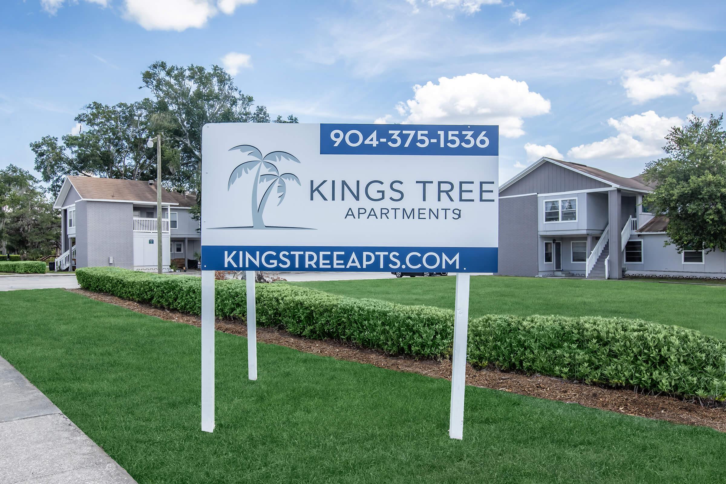 Sign for Kings Tree Apartments featuring a palm tree graphic. Contact number: 904-375-1536. Website: kingstreeapts.com. The background shows well-maintained green lawns and residential buildings.