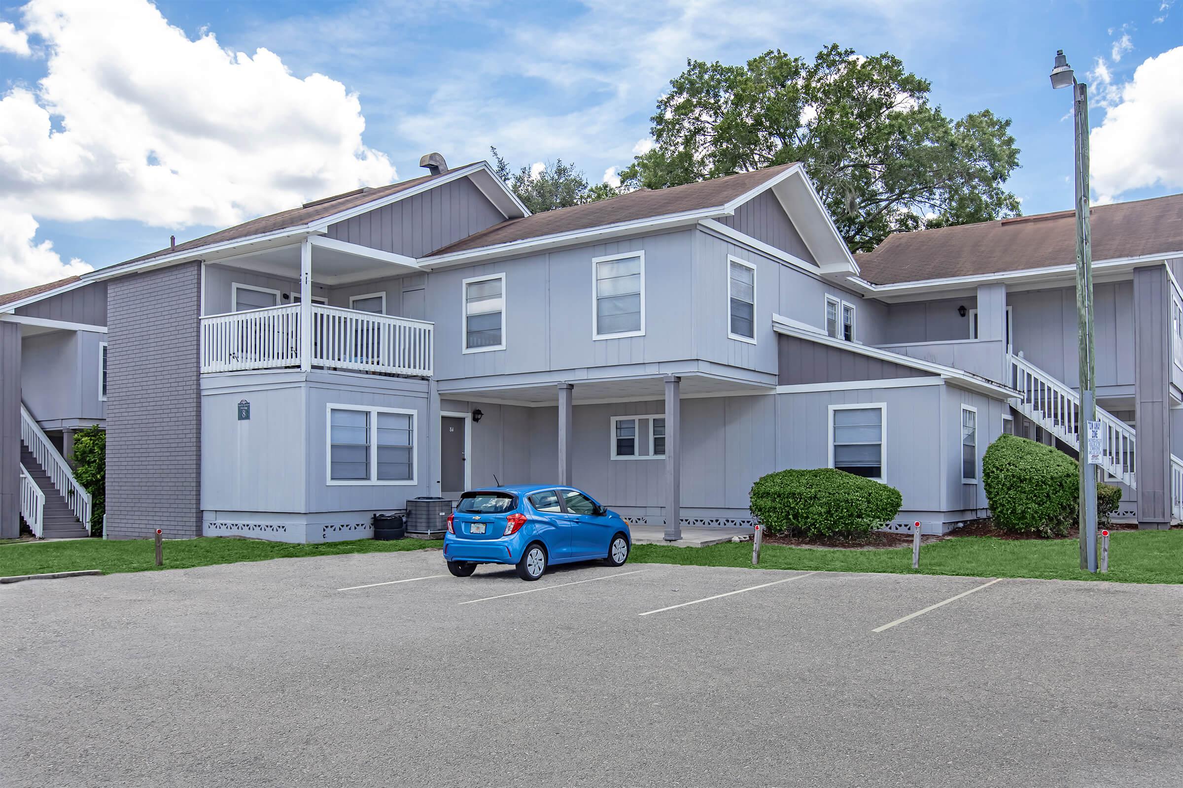 A two-story residential building with a combination of light gray and white siding, featuring balconies and stairs. In the foreground, a blue car is parked in a lot with several empty spaces. There are green shrubs and trees surrounding the building, with a partly cloudy sky overhead.