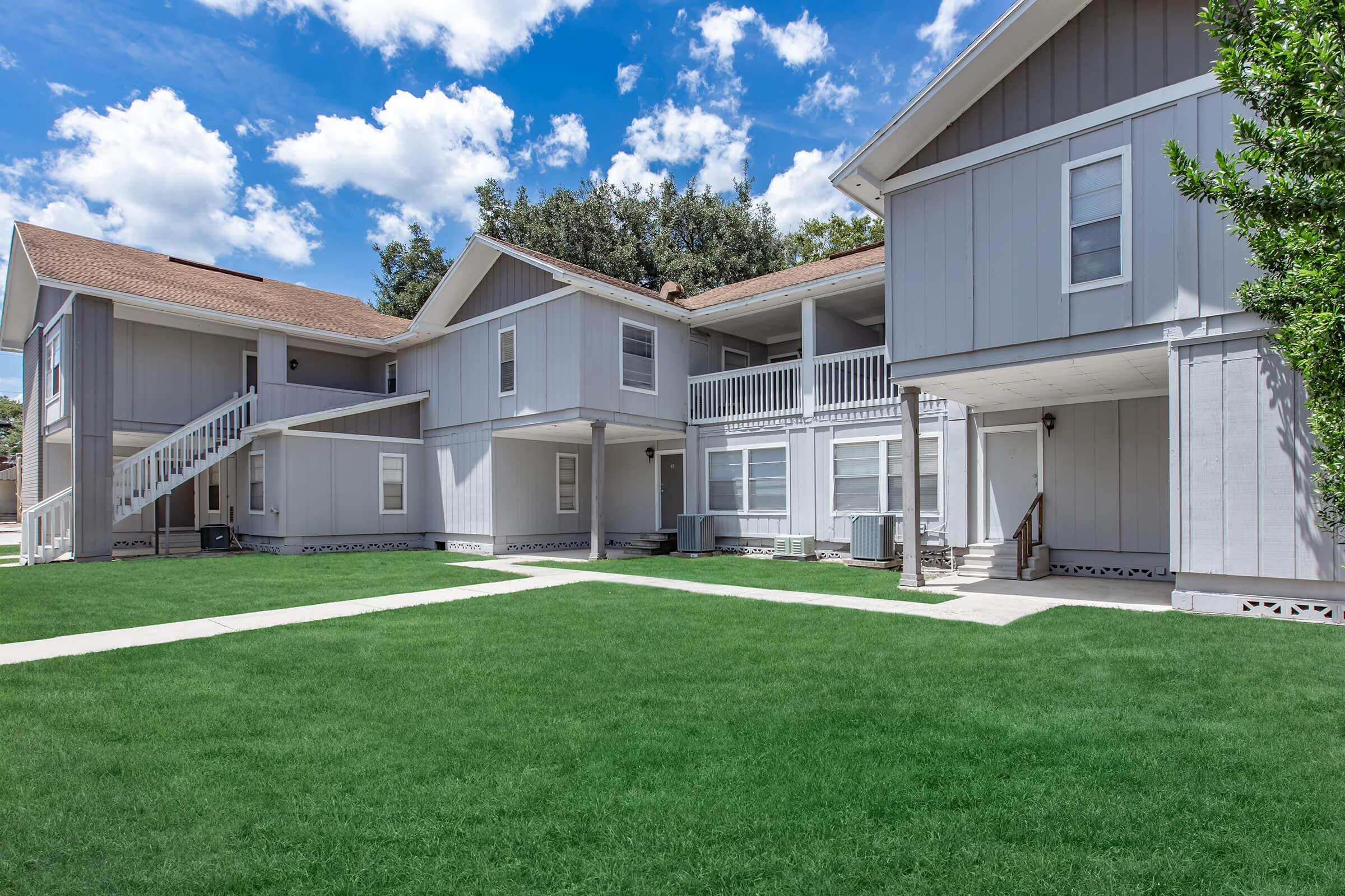A well-maintained multi-unit residential building with gray siding, featuring a green lawn, white stairs, and clear blue skies with scattered clouds.