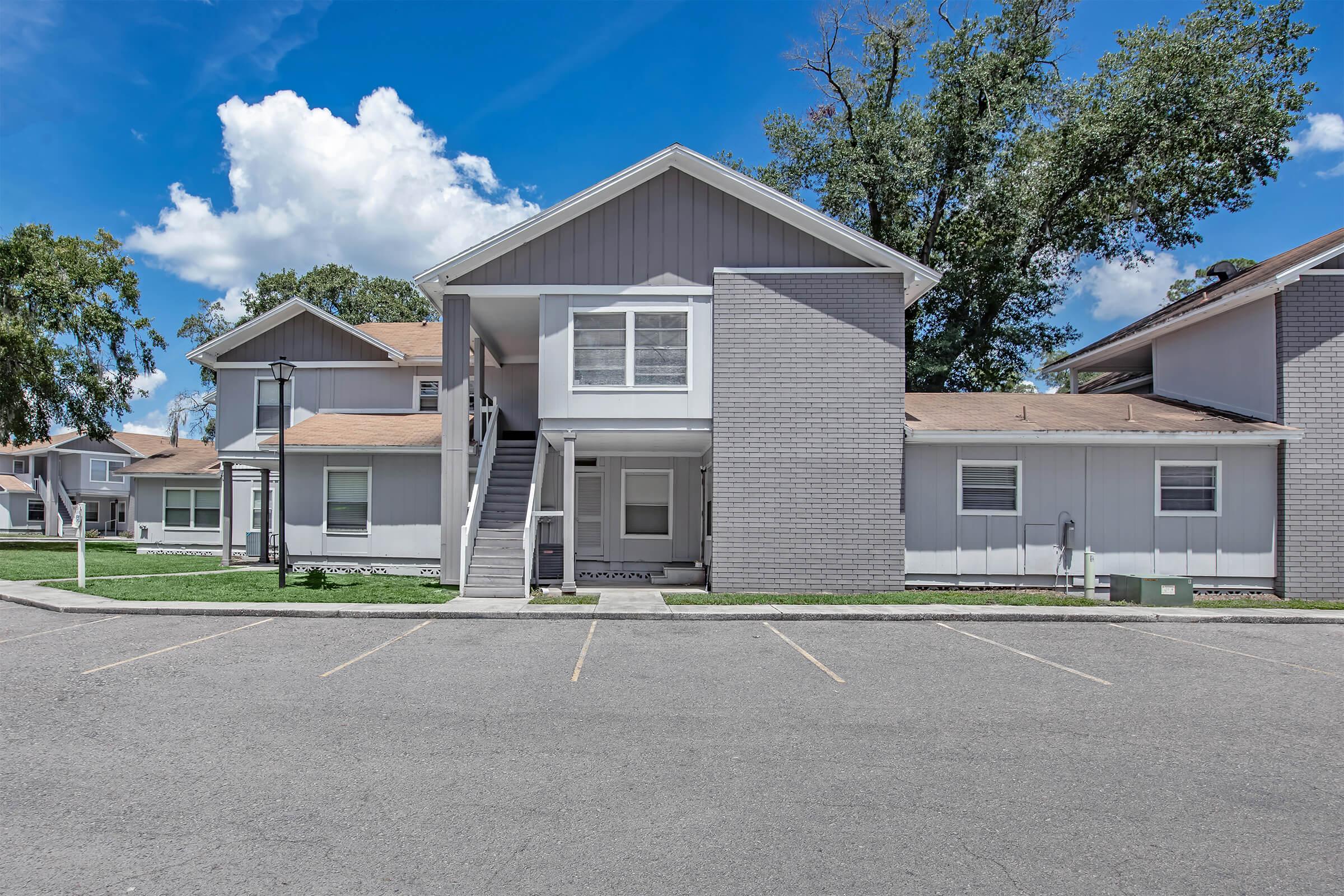 A multi-unit residential building featuring grey and white exteriors with a staircase leading to the upper level. The setting includes a paved parking area and green grass, under a clear blue sky with a few clouds. The scene conveys a well-maintained apartment complex.
