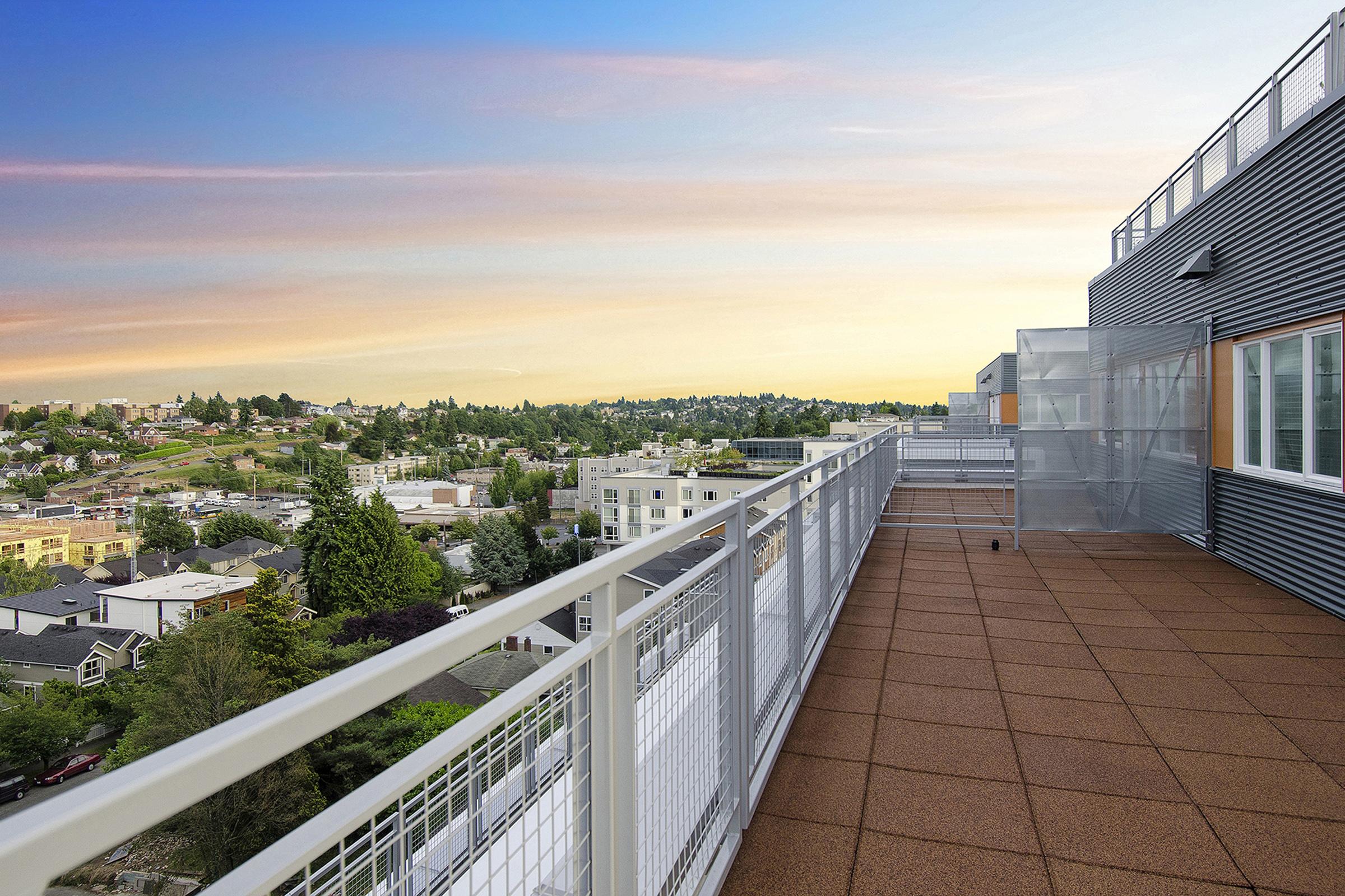 A spacious balcony overlooking a cityscape at sunset, featuring a railing and modern building design. The scene includes greenery and a variety of residential buildings in the background, enhancing the view and ambiance. The sky is painted with soft pastel colors, creating a serene atmosphere.