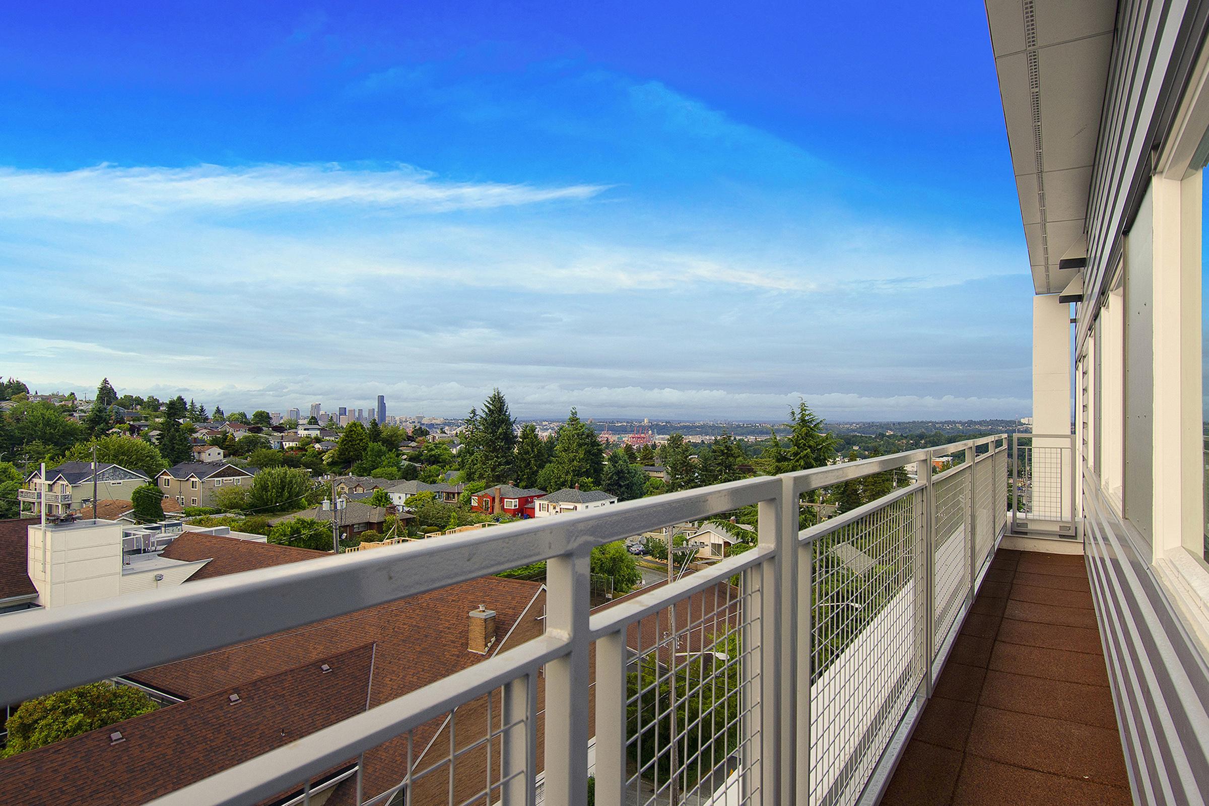 View from a balcony overlooking a cityscape, featuring residential neighborhoods and greenery under a clear blue sky with scattered clouds. The railing of the balcony is visible in the foreground, emphasizing the height and perspective of the vantage point.