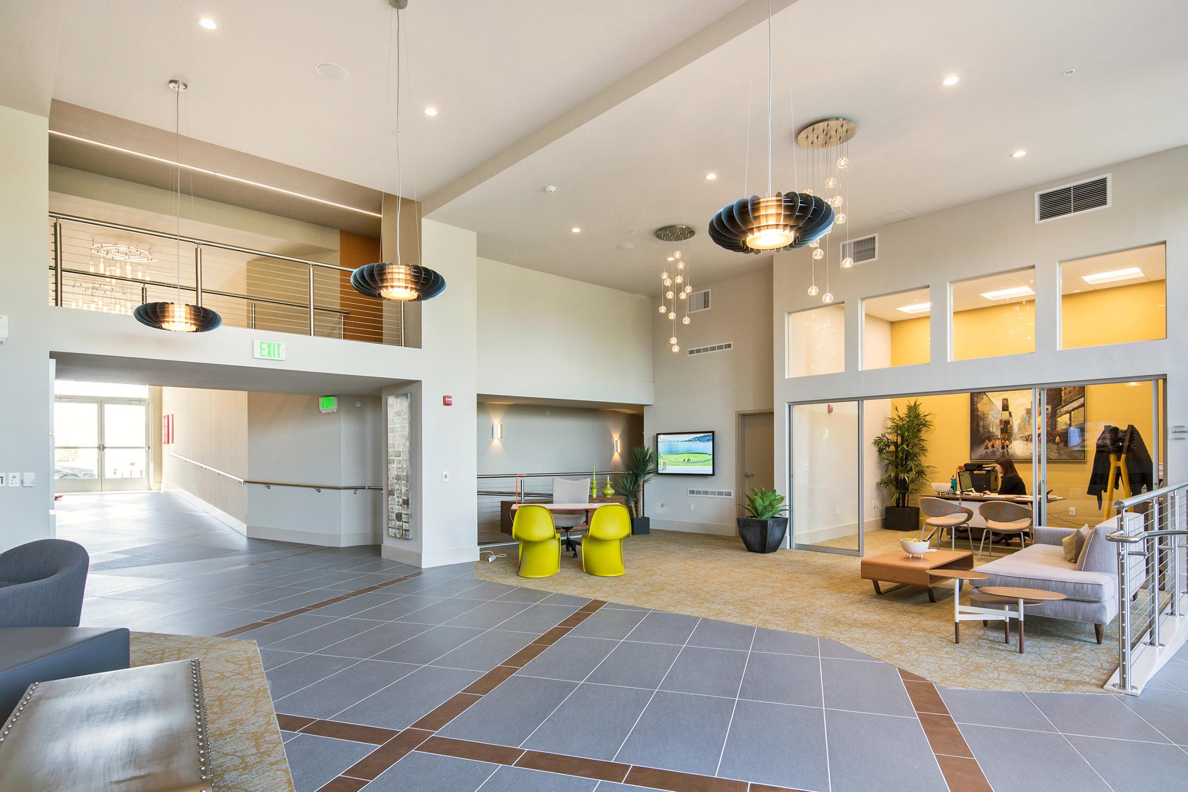 A spacious modern lobby featuring stylish furniture, large windows, and contemporary lighting. The area includes a reception desk, seating arrangements with plush chairs, decorative plants, and a TV screen. The floor has a mix of tile and carpet, creating an inviting atmosphere.