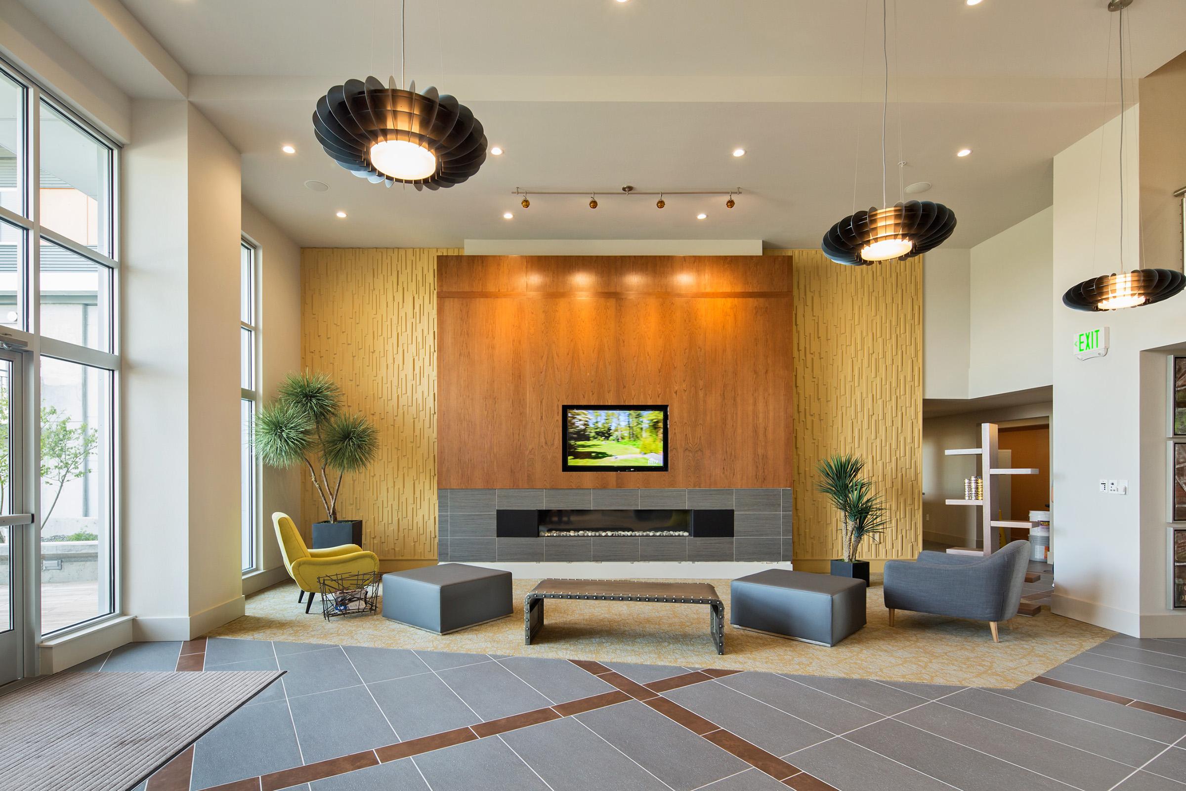 A modern lobby featuring a wooden accent wall with a TV, surrounded by stylish furniture including a yellow chair, gray sofa, and a bench. Tall indoor plants add greenery, while hanging pendant lights provide illumination. The floor has a combination of tiles and carpet.