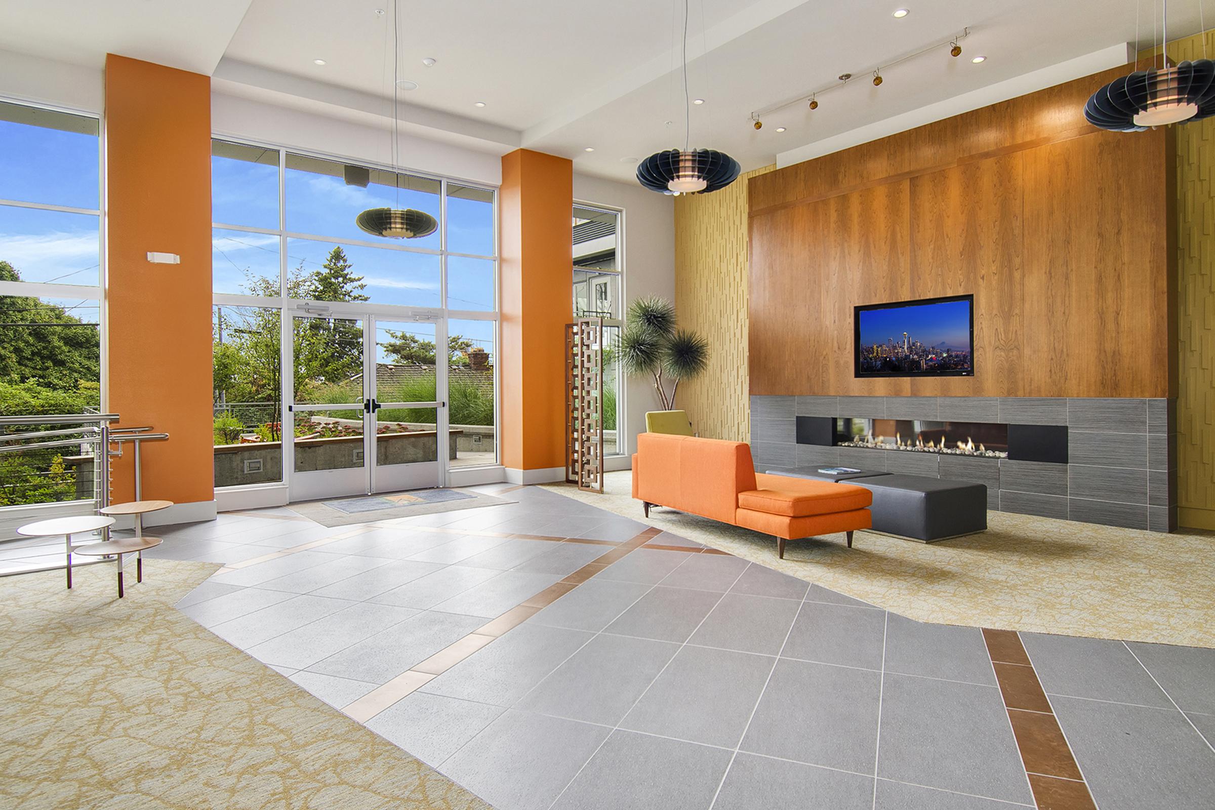 A modern lounge area with large windows allowing natural light. The walls are painted in orange and neutral tones. An orange sofa is positioned near a wall-mounted TV, and there’s a contemporary fireplace below it. Stylish light fixtures hang from the ceiling, and plants add a touch of greenery.