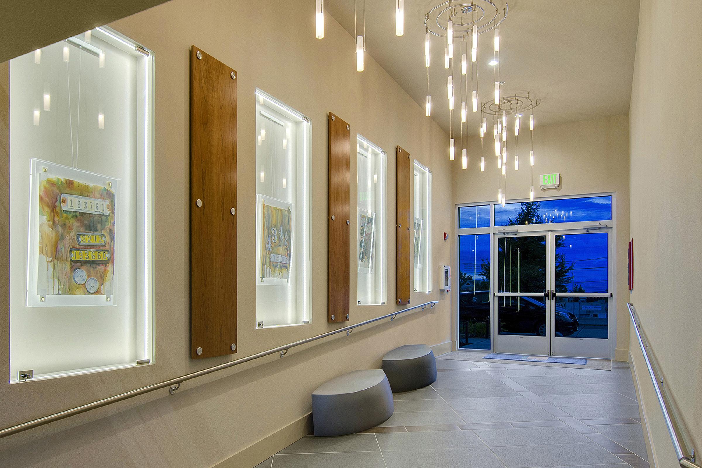 A modern, well-lit hallway featuring framed art pieces displayed in backlit cases along the walls. The space has a sleek design with wooden panels, contemporary lighting fixtures hanging from the ceiling, and a glass door at the end leading to the outside. Two rounded gray seating areas are positioned in the corridor.