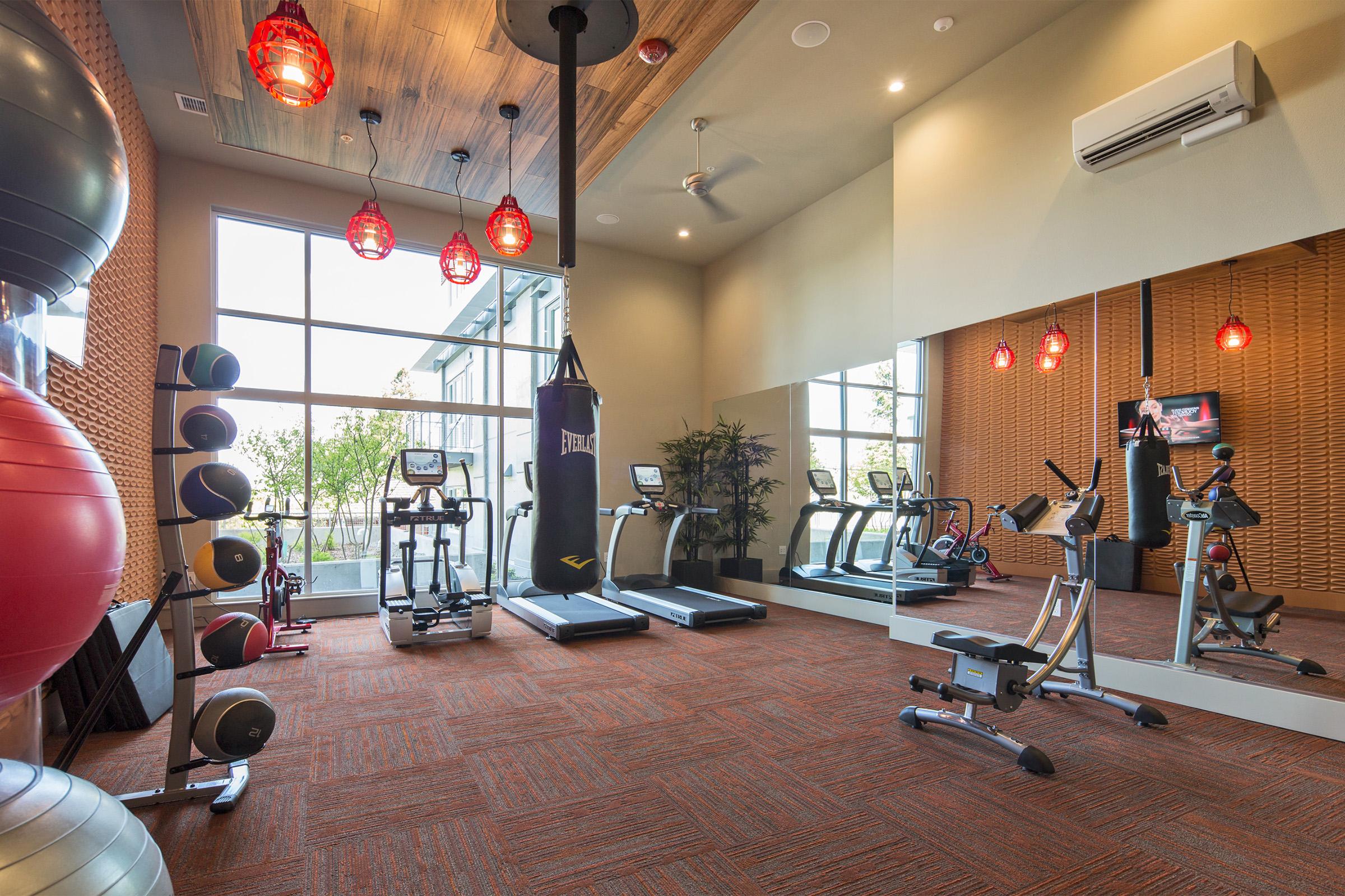 A bright and modern fitness room featuring exercise equipment such as treadmills, a stationary bike, and free weights. Large windows provide natural light, and decorative red pendant lights hang from the ceiling. A mirror runs along one wall, enhancing the sense of space.
