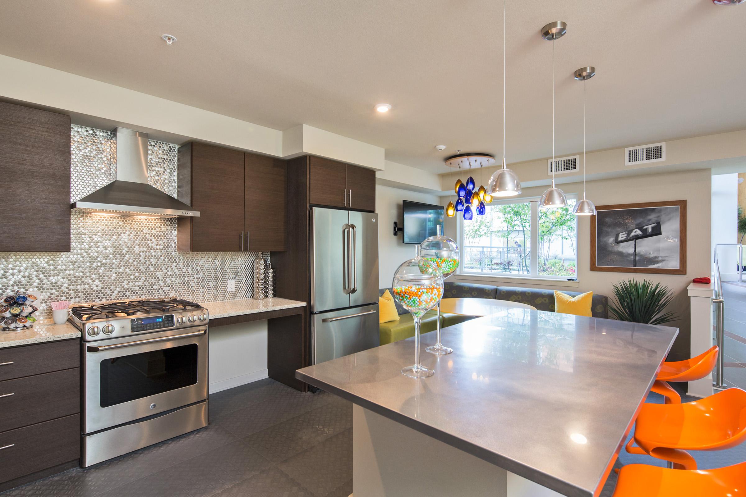 Modern kitchen featuring a stainless steel stove and refrigerator, dark wood cabinets, and a stylish backsplash. A central island with a concrete countertop and colorful bar stools is visible. Large windows provide natural light, creating a bright, inviting atmosphere. A decorative glass bowl adds a pop of color.
