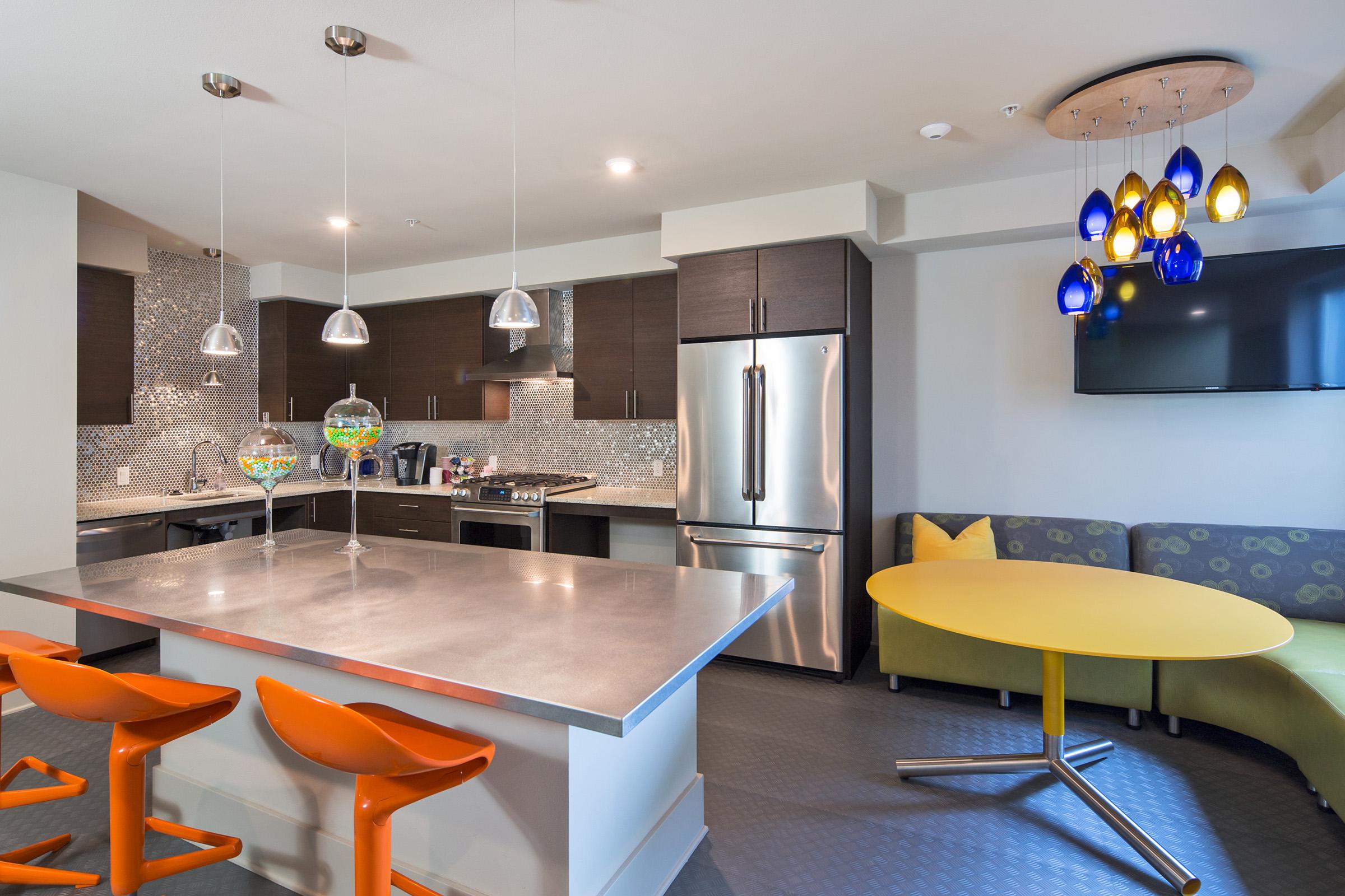 Modern kitchen featuring stainless steel appliances, sleek cabinetry, and a large island with orange bar stools. A round yellow dining table is situated next to a curved gray sofa with a decorative pillow. Unique pendant lights and a TV mounted on the wall add to the contemporary design.