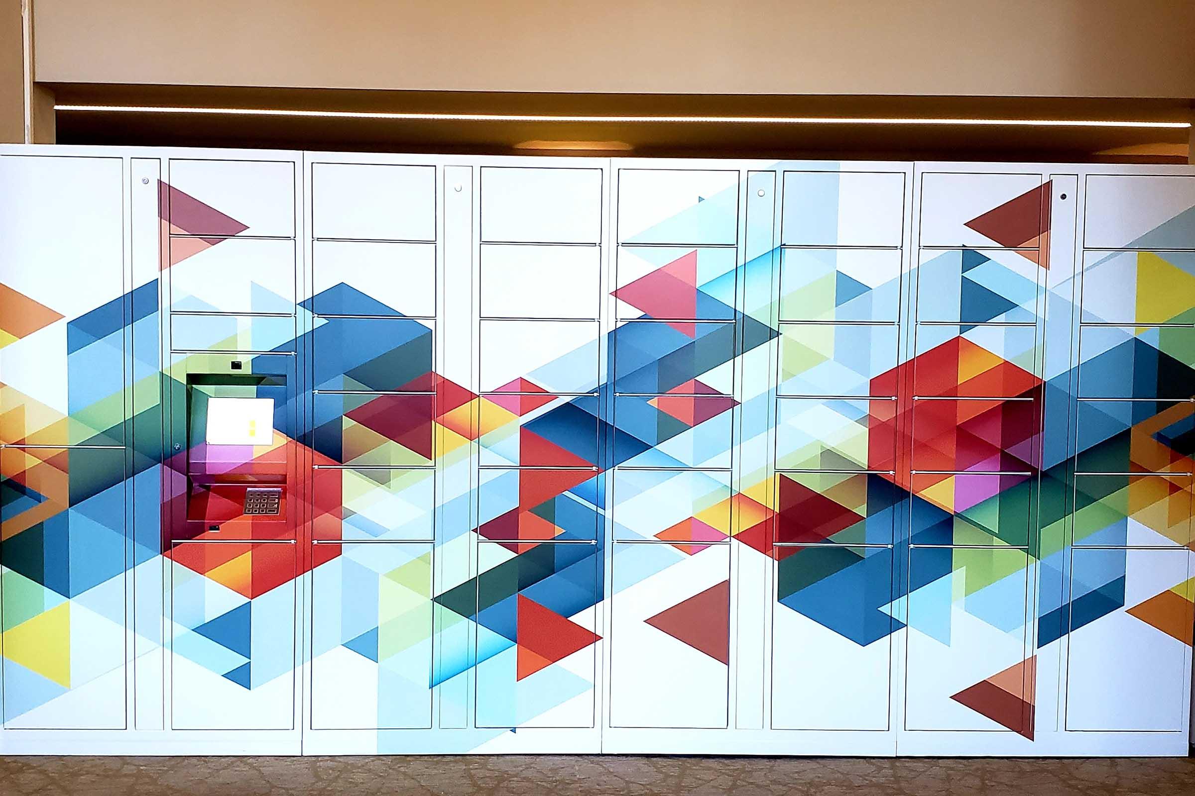 A row of colorful lockers featuring a geometric design with various shades of red, blue, green, and yellow. One locker has an electronic keypad and screen, suggesting it may be used for package deliveries or secure storage. The overall aesthetic is modern and vibrant.