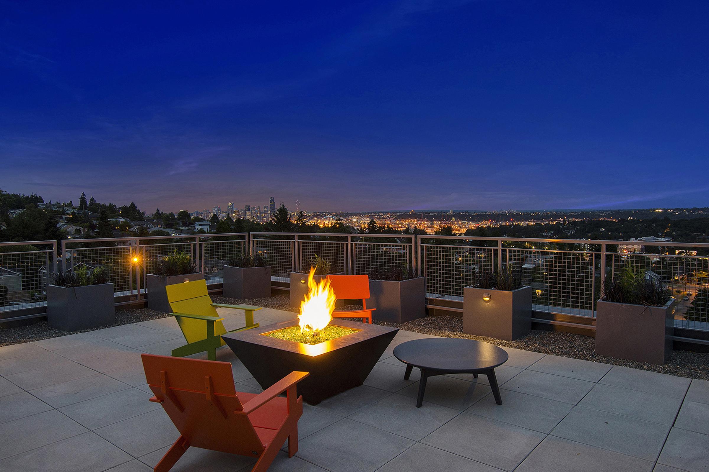 A rooftop patio at dusk featuring a modern fire pit surrounded by colorful chairs. The view overlooks a city skyline illuminated by lights against a twilight sky. Planters with greenery line the edge of the patio, adding a touch of nature to the urban setting.