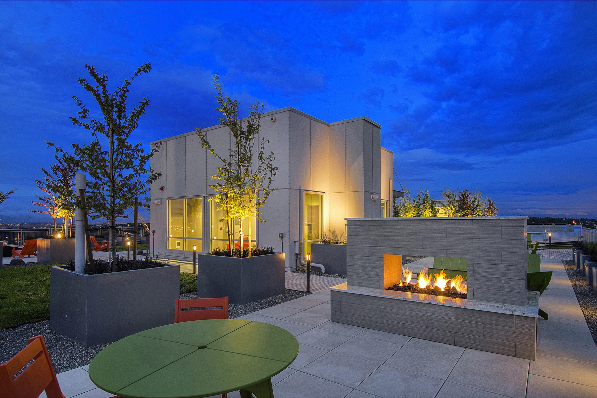 Modern rooftop space featuring a cozy fire pit surrounded by plants. In the background, a sleek building with large windows is illuminated under a twilight sky. Colorful seating arrangements are visible, enhancing the inviting atmosphere of the outdoor area.
