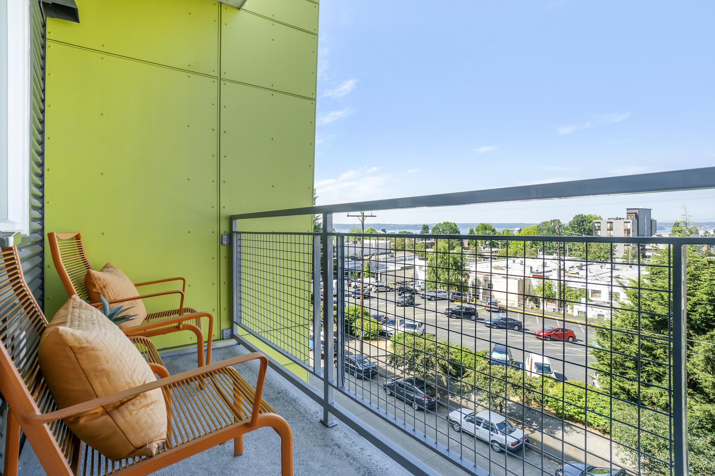 A balcony with two orange chairs and pillows, overlooking a street and green trees. The building features a bright green exterior wall, with a clear blue sky in the background.