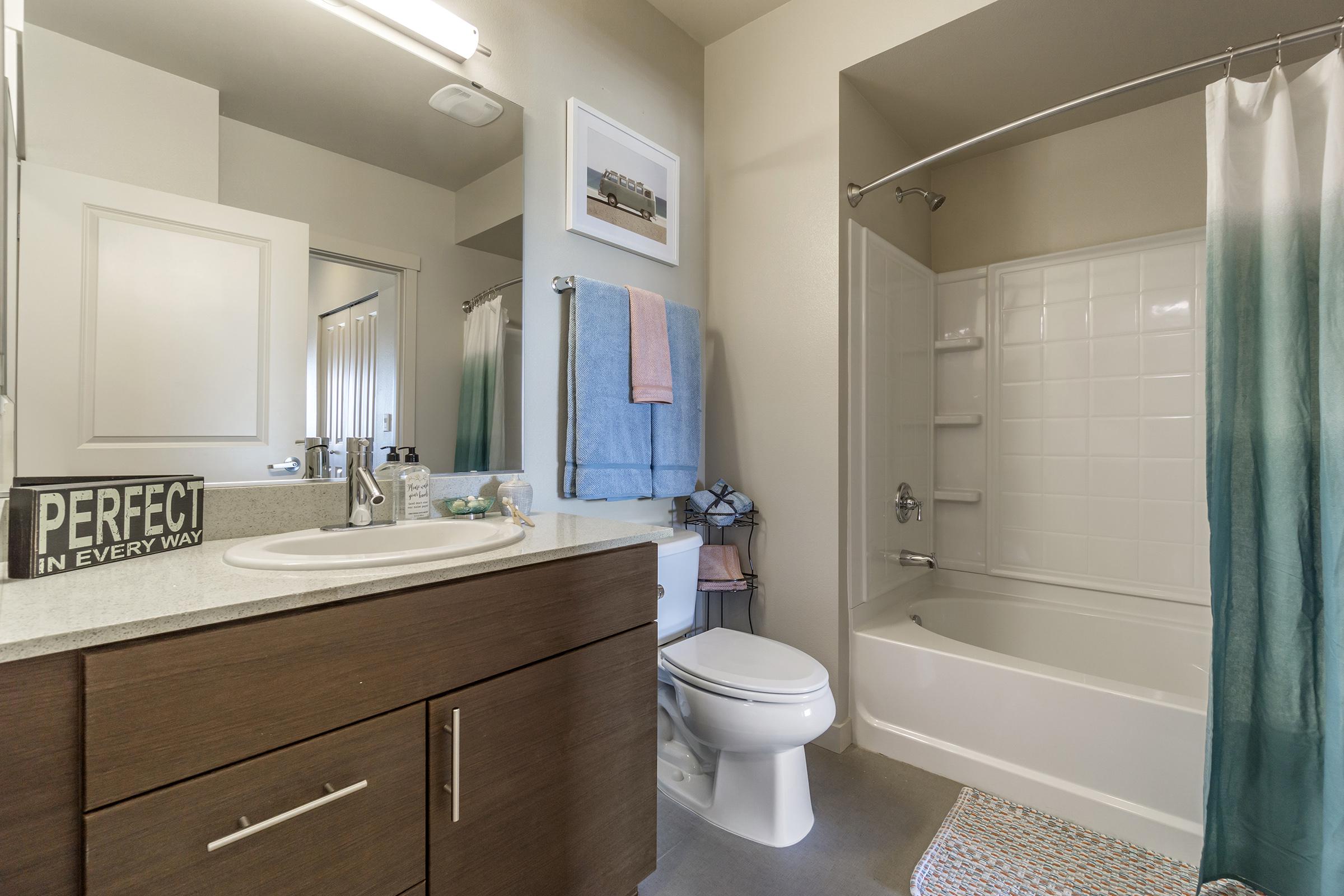 A modern bathroom featuring a white bathtub with a shower, a sink with dark wood cabinetry, and a large mirror. There are towels hanging on a rack and a decorative sign on the countertop. The color scheme includes soft neutrals and light blue accents. Natural light illuminates the space.
