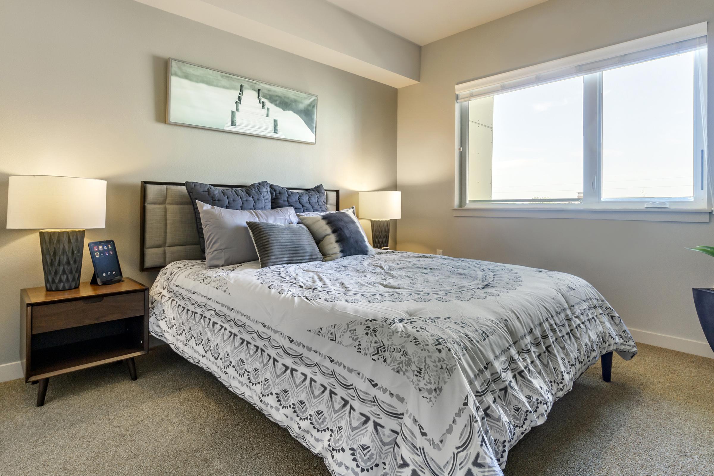 A cozy bedroom featuring a neatly made bed with decorative pillows, a stylish light-colored bedspread, and two bedside lamps on either side of a wooden nightstand. A modern piece of artwork hangs above the bed, and a large window allows natural light to fill the space.