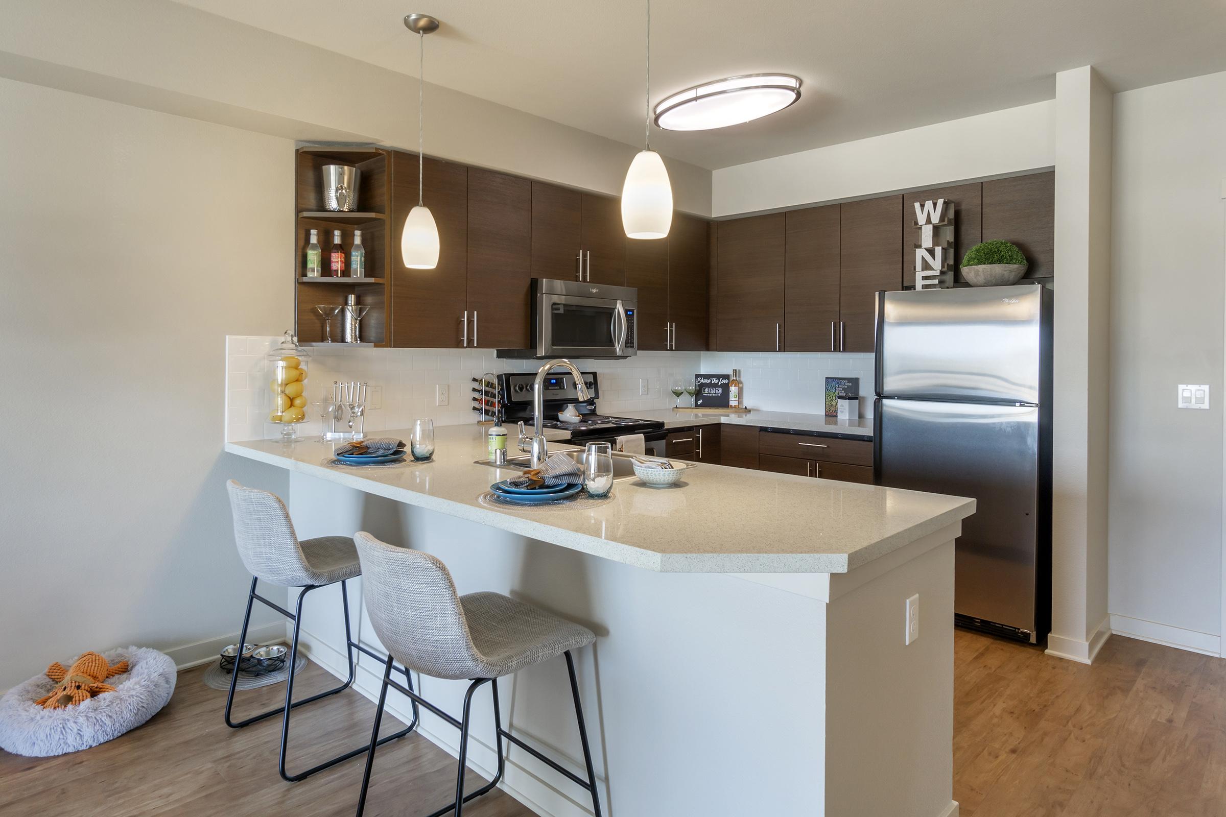 Modern kitchen featuring sleek cabinetry, stainless steel appliances, and a spacious island with bar seating. Pendant lights hang above the island, and decorative items are arranged on shelves. A cozy pet bed is visible on the floor, adding a touch of homey comfort.