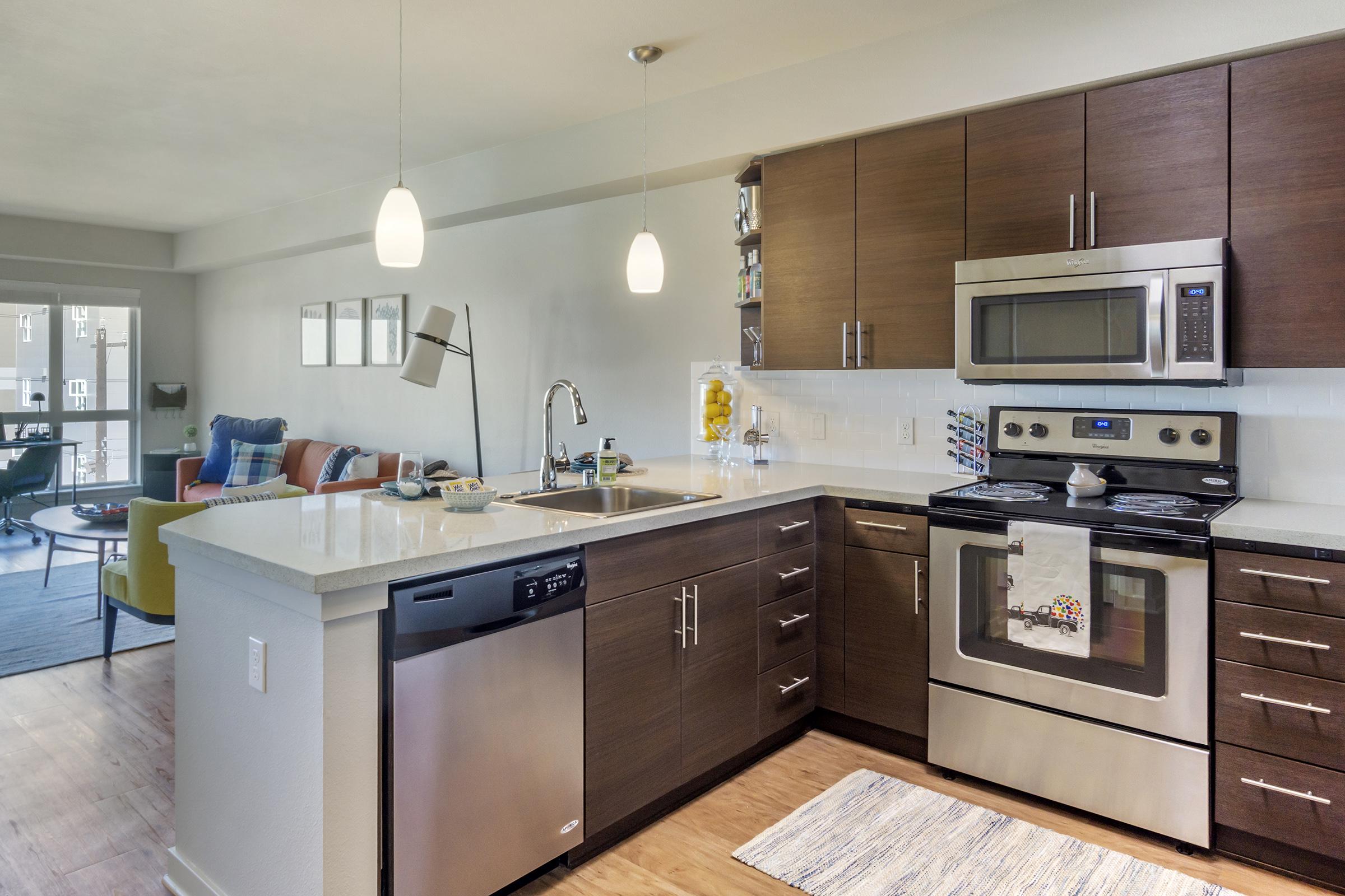 Modern kitchen featuring stainless steel appliances, including a microwave, oven, and dishwasher. The space has dark wood cabinetry and a white countertop. A cozy living area is visible in the background with colorful furniture and large windows, enhancing the open layout and natural light.