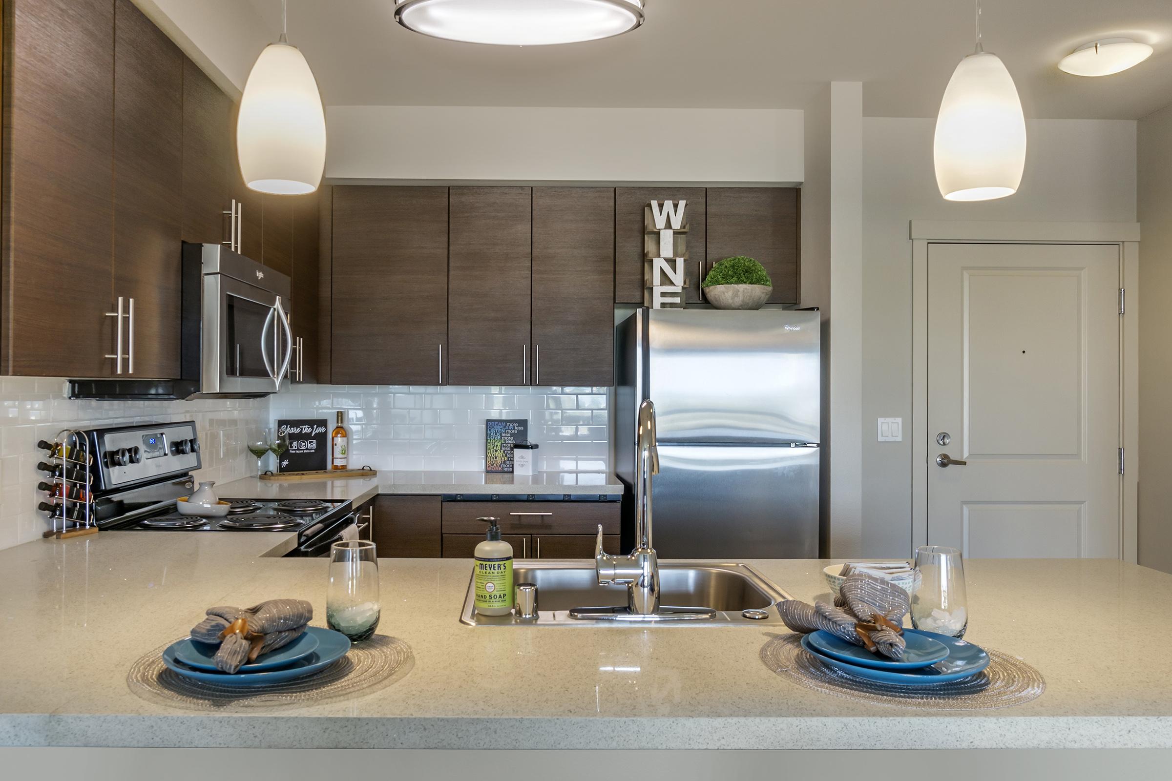 A modern kitchen featuring dark wood cabinets, stainless steel appliances, and a spacious countertop. The island is set with decorative plates and glasses, while pendant lights illuminate the space. A small plant and a sign that says "WINE" add personalized touches.