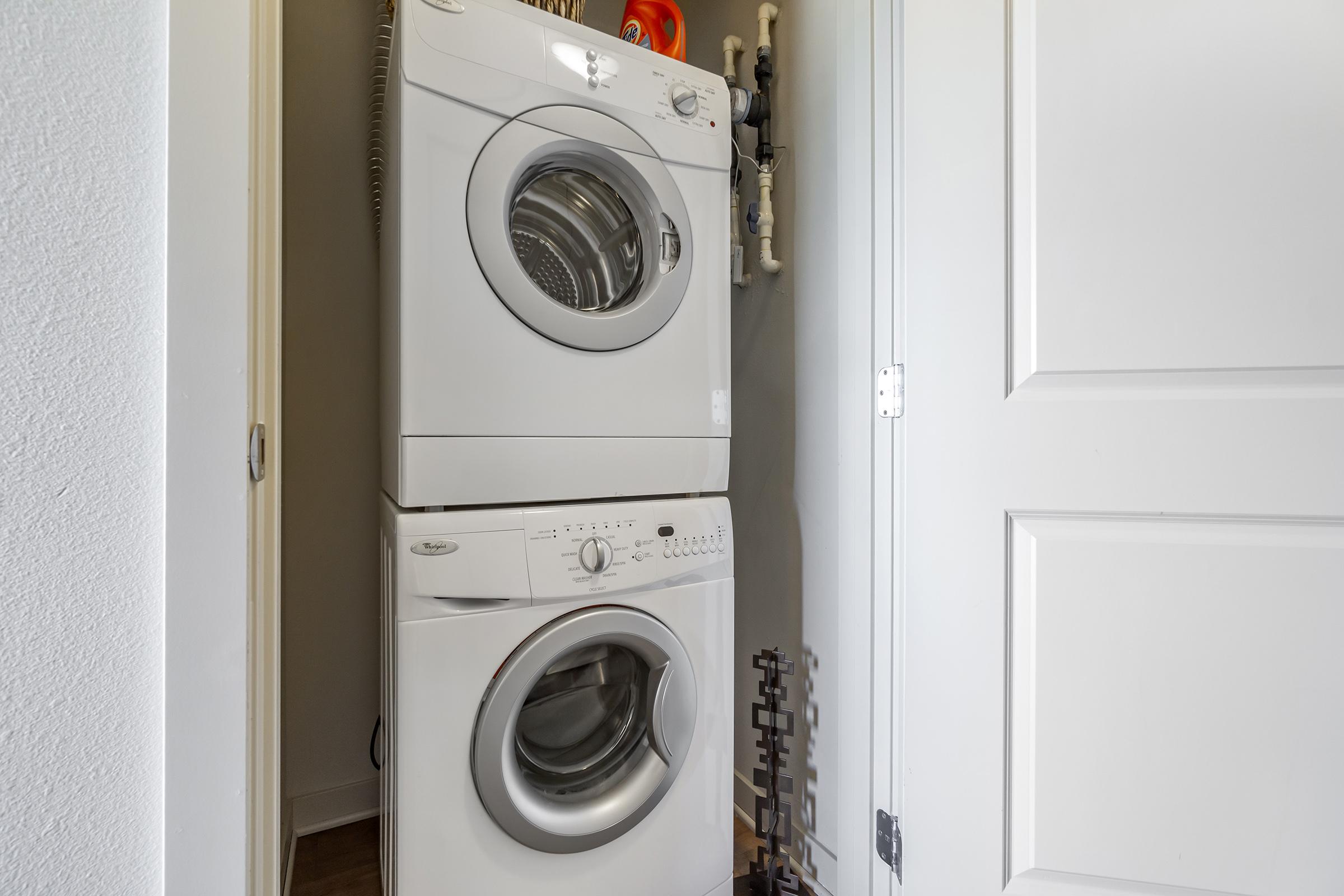 A stackable washer and dryer unit in a small laundry area, positioned near a door. The machines are white and appear modern, with controls on the front of the dryer. The space is well-lit, featuring light-colored walls and a glimpse of plumbing behind the units.