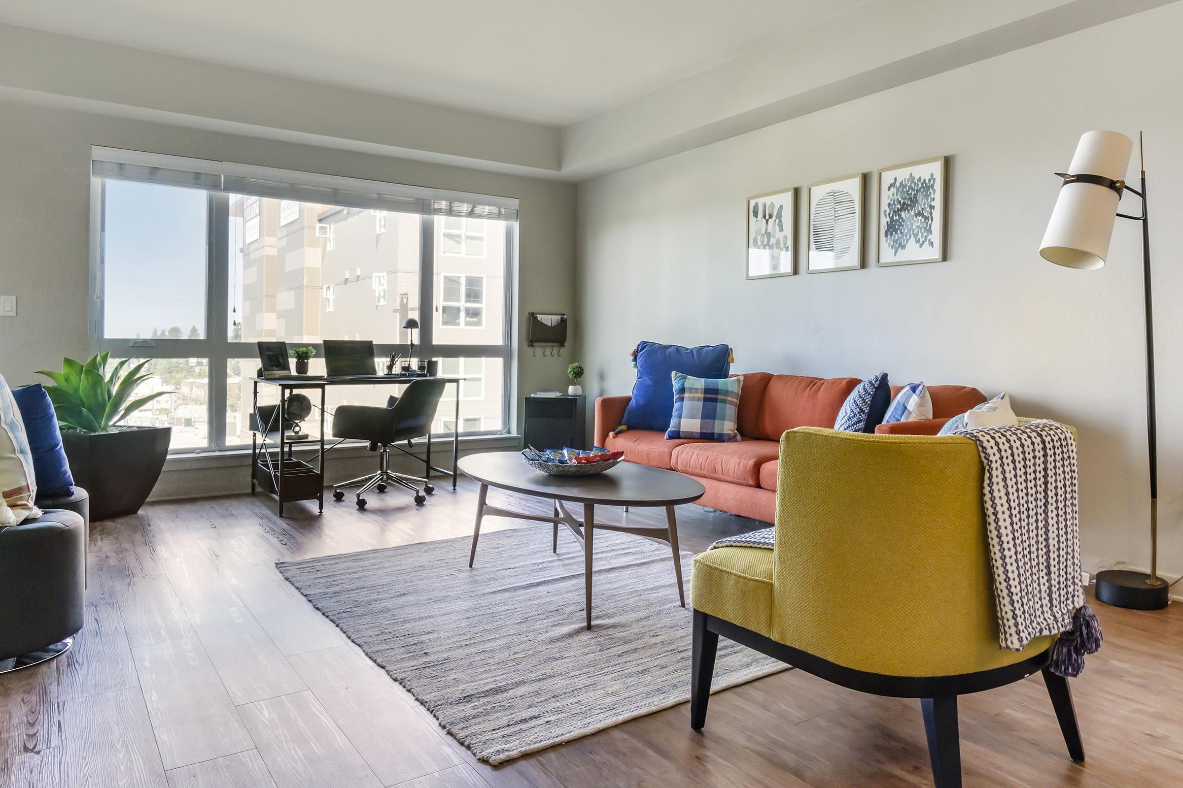 A modern living room featuring a large window with city views, a coral sofa with blue and white cushions, a yellow armchair draped with a blanket, a round coffee table, and a workspace with a desk and chair. Decorative plants and framed artwork add a touch of style to the space.