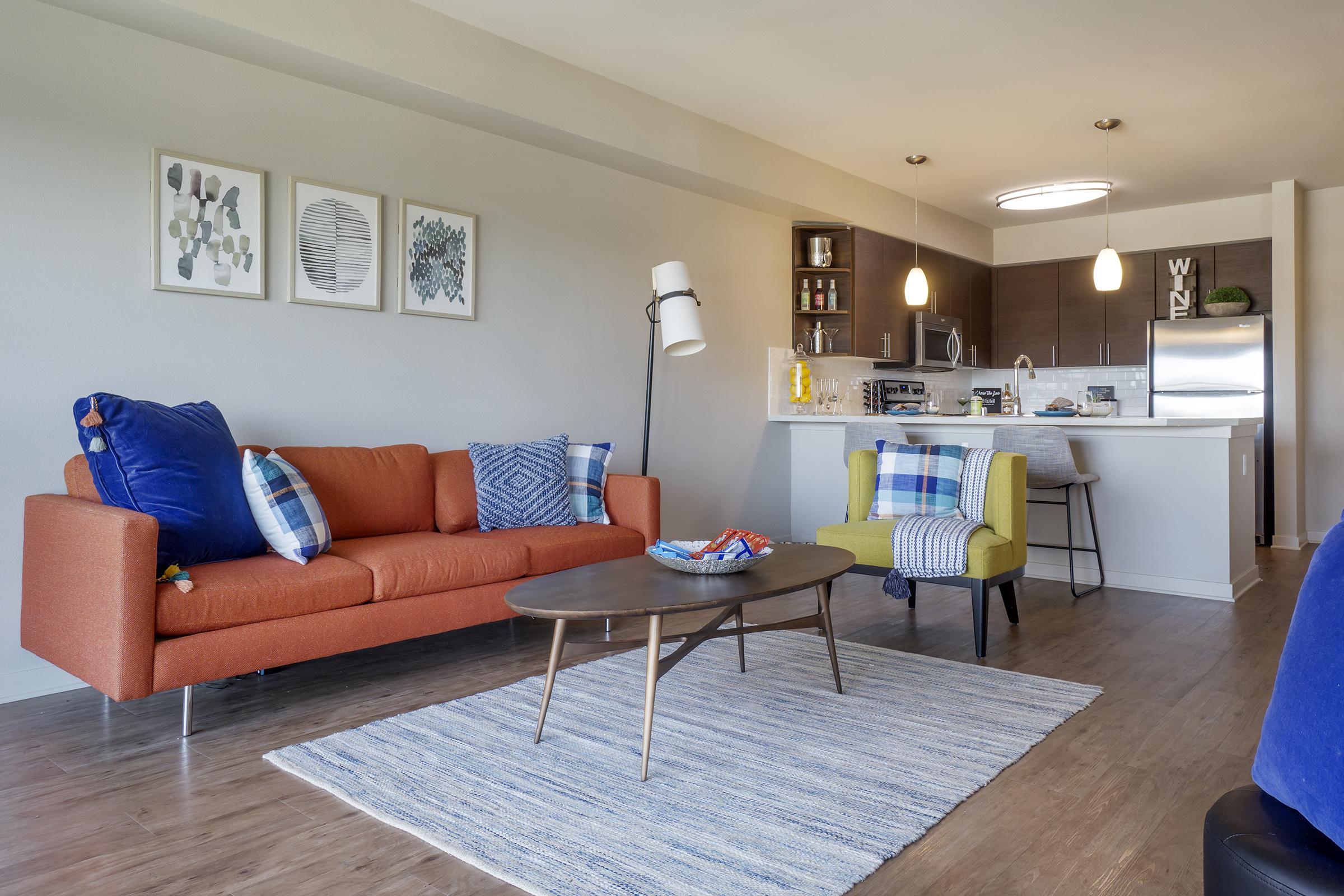 A stylish living room featuring an orange sofa with blue and patterned pillows, a round coffee table, and a light area rug. A kitchenette with modern appliances is visible in the background, illuminated by pendant lighting. Wall art adds a decorative touch to the space, creating a cozy atmosphere.