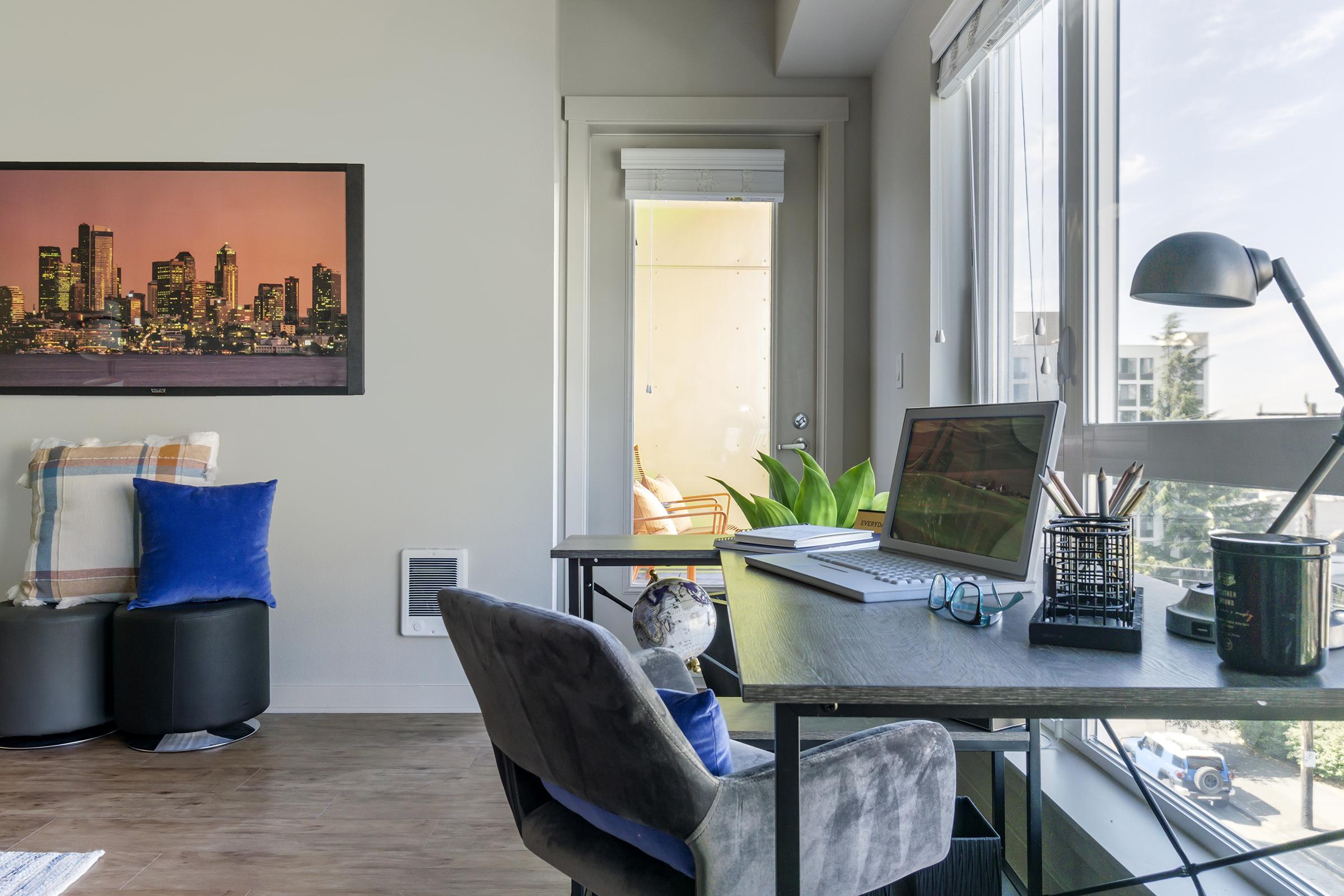 A modern home office space featuring a desk with a laptop, stationery, and a small globe. Directly outside the window, a view of a city skyline at sunset is visible. The room has a cozy lounge area with a chair and potted plants, and warm, natural lighting enhances the inviting atmosphere.