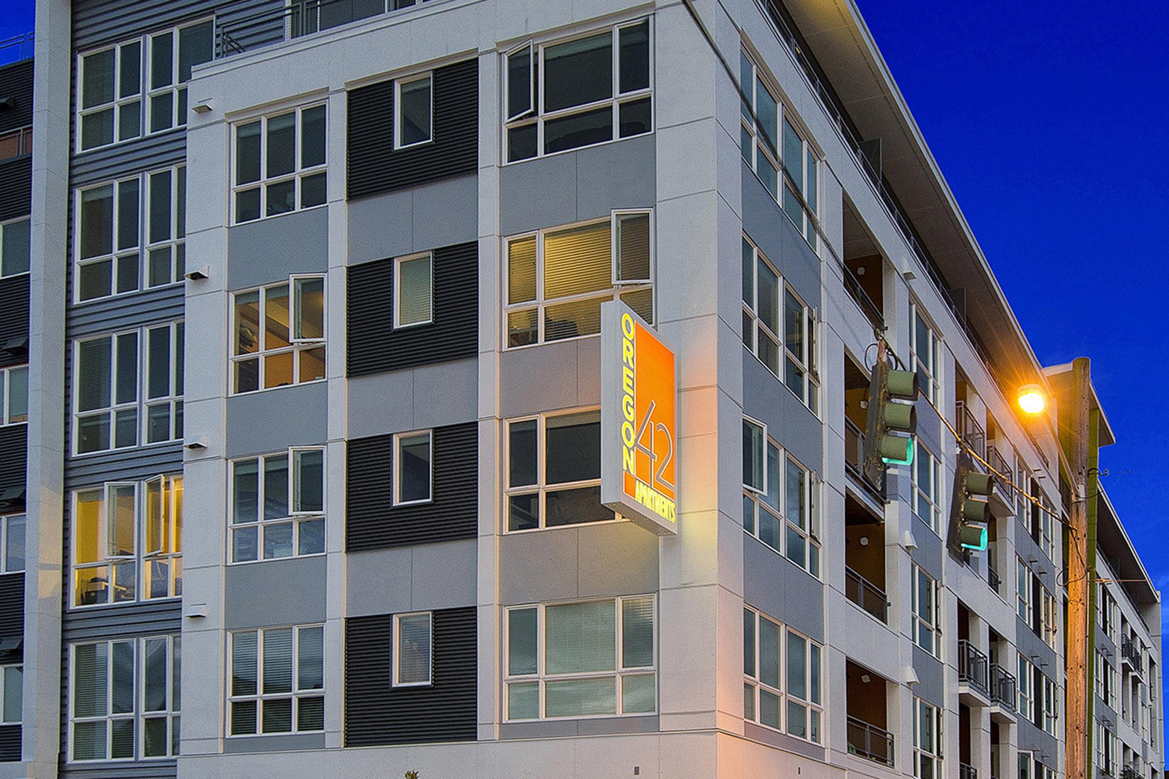 Modern apartment building with multiple windows, featuring a prominent illuminated sign that reads "Oregon 12." The building has a mix of light and dark gray panels, and there's street lighting visible in the foreground against a twilight sky.