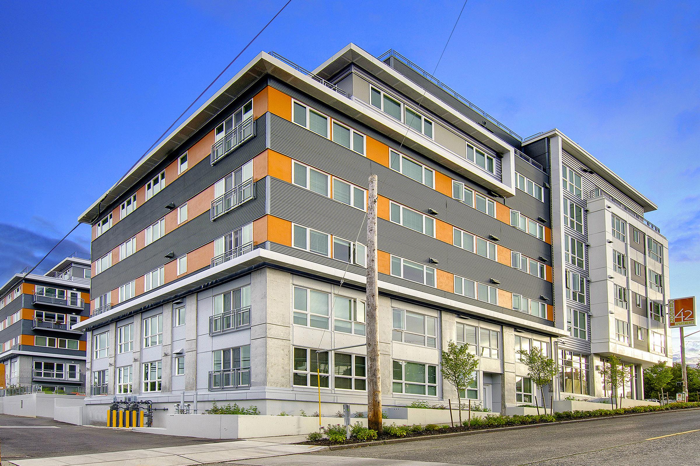 Modern apartment building featuring multiple stories with a mix of gray and orange exterior panels. Large windows and a sleek design are prominent, along with well-maintained landscaping and a sidewalk. The blue sky adds a vibrant backdrop to the architecture.