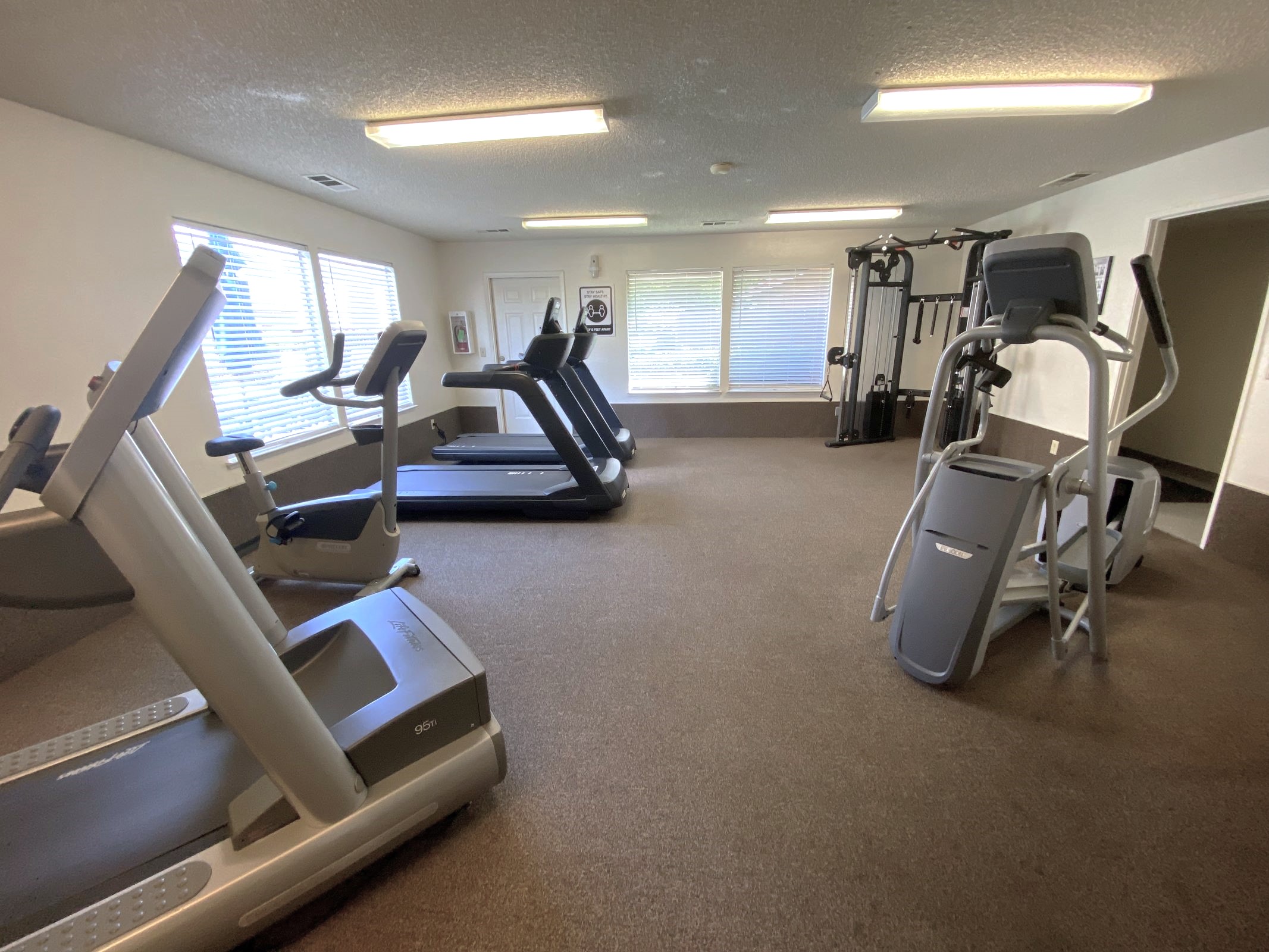A spacious, well-lit gym with various exercise equipment, including treadmills, an elliptical machine, and a strength training station. Large windows let in natural light, and the room features a carpeted floor, creating a welcoming environment for workouts.