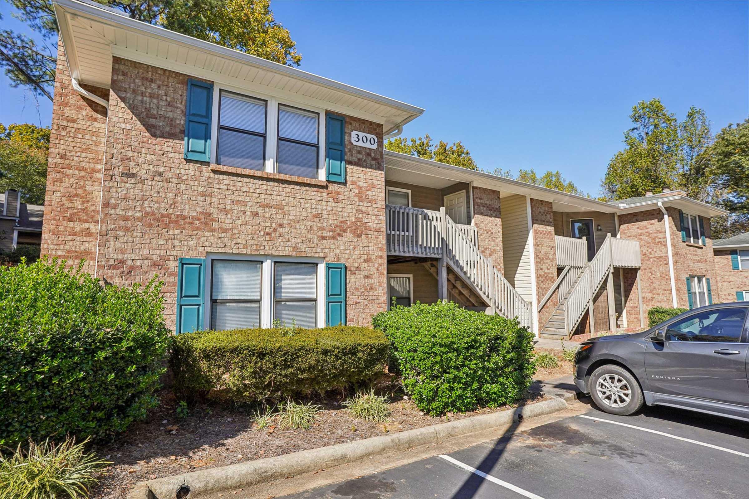 A two-story brick apartment building with blue shutters and a staircase leading to the second floor. The building is surrounded by well-maintained shrubs and small trees, with a parking area in front. The number "300" is visible on the exterior.