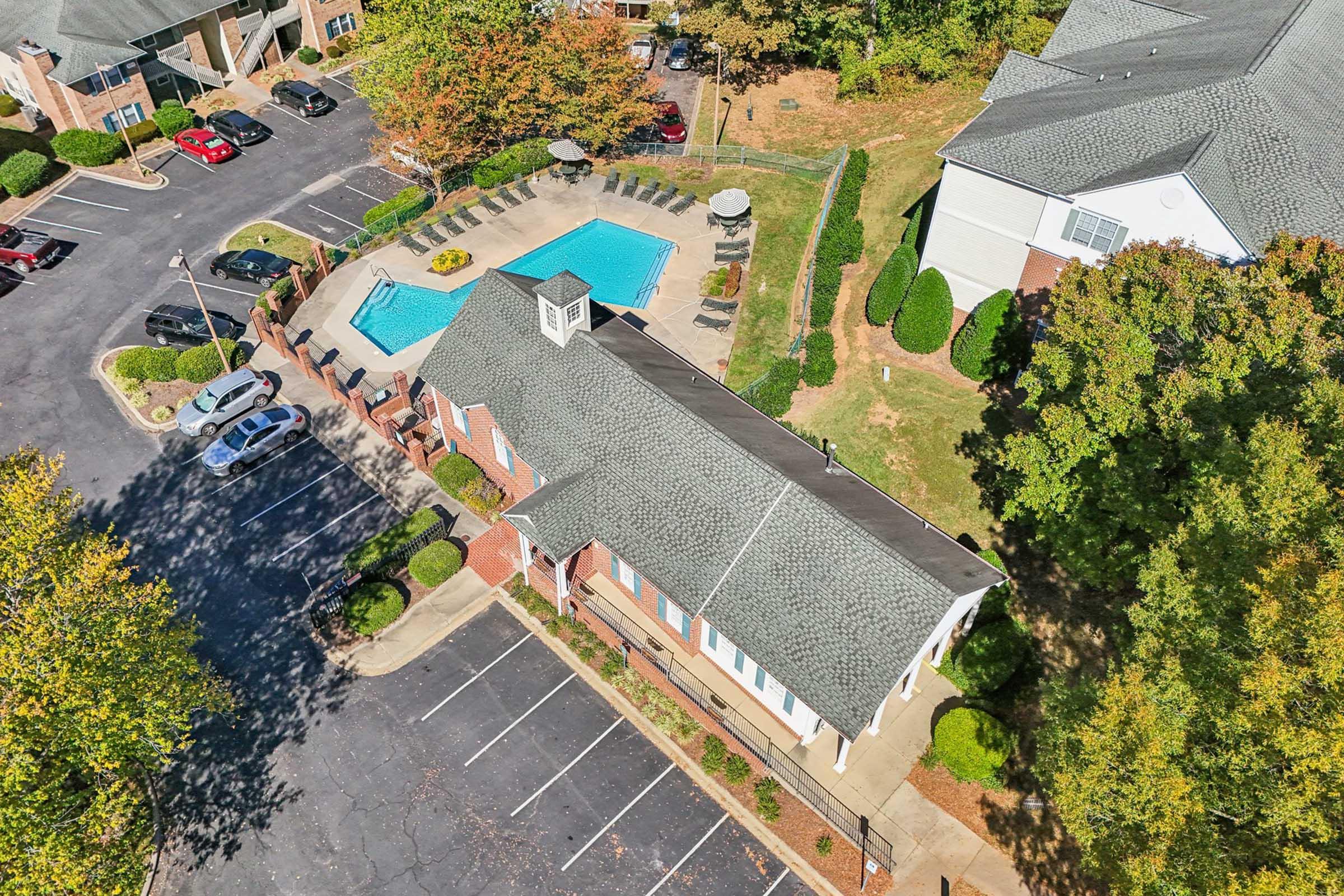 Aerial view of an apartment complex featuring a swimming pool surrounded by lounge chairs. Nearby cars are parked in an organized layout. The building has a sloped roof and is situated among trees with autumn-colored leaves.