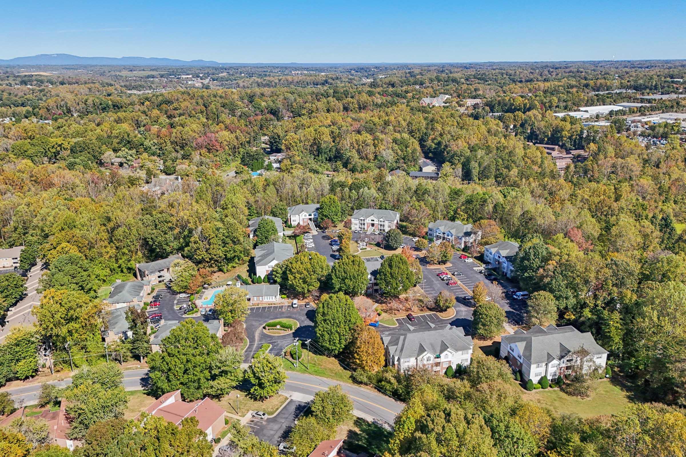 Aerial view of a suburban neighborhood surrounded by trees showcasing various residential buildings and parked cars. Spacious green areas and winding roads can be seen, with a mix of single-family homes and apartment complexes amid vibrant fall foliage. Blue sky and distant mountains complete the serene landscape.