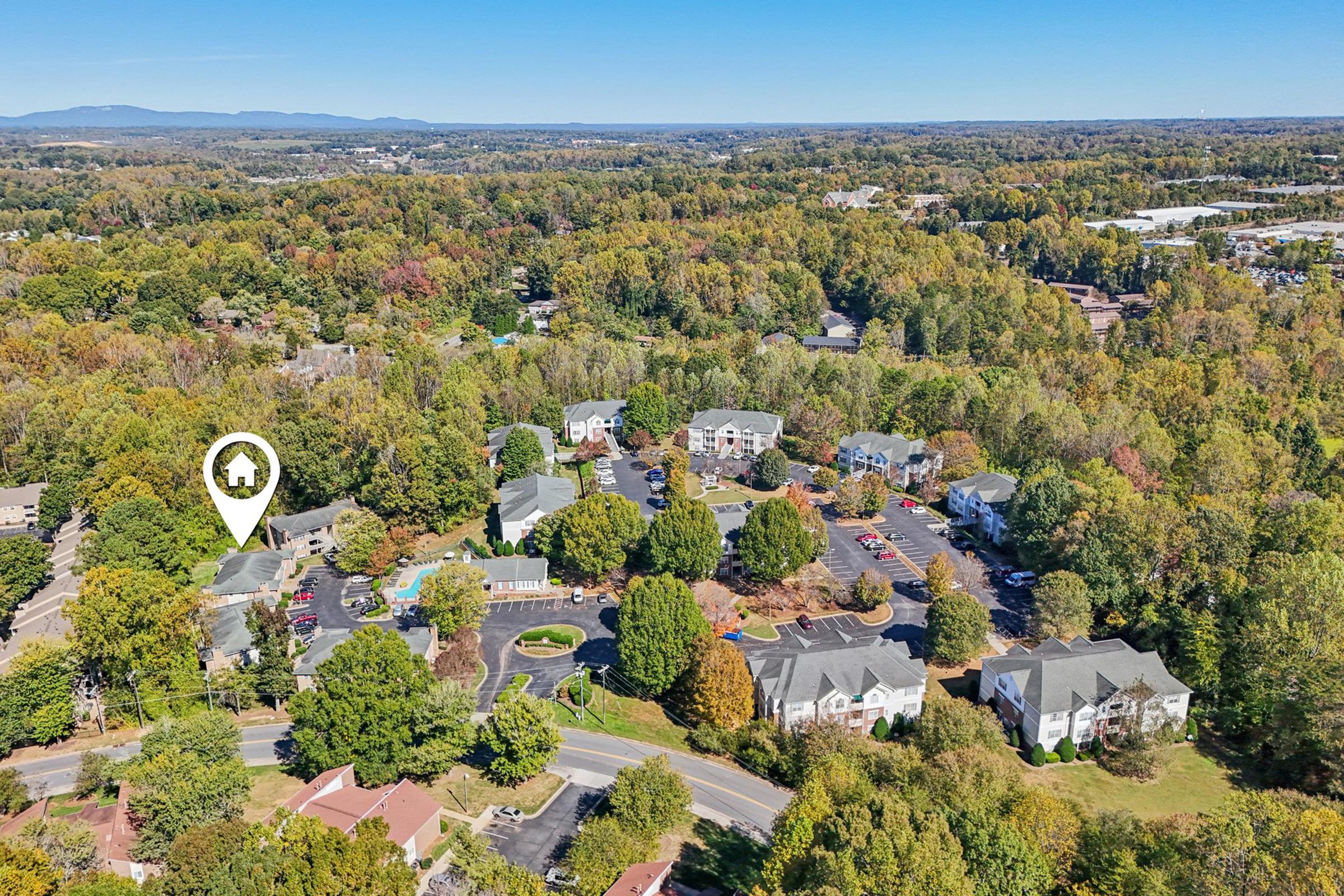 An aerial view of a residential community surrounded by trees, showcasing various buildings and green spaces. A designated location marker is visible, indicating a specific point of interest within the neighborhood. The landscape features a mix of residential units and open areas, with distant mountains on the horizon.