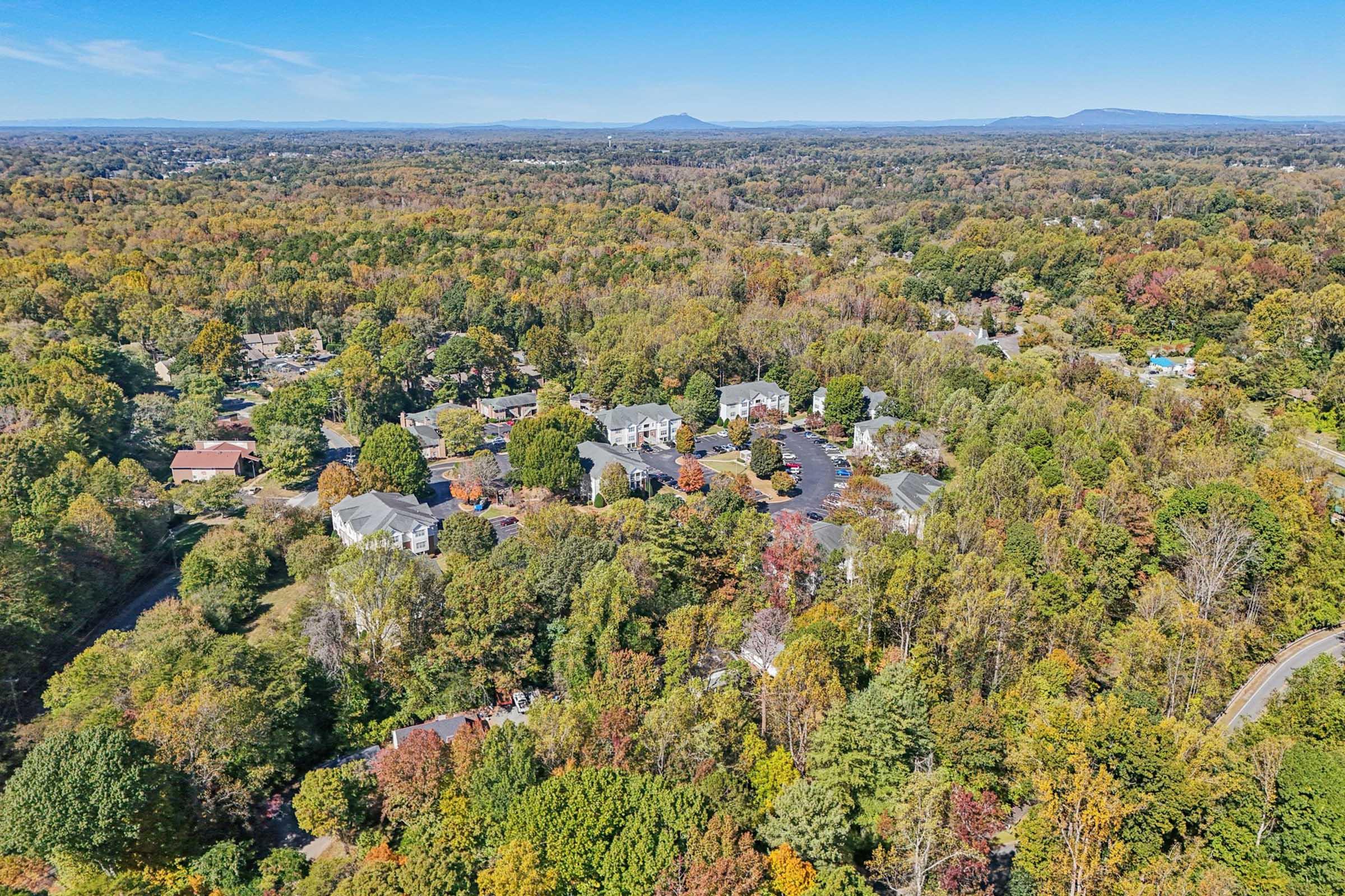 Aerial view of a colorful autumn landscape featuring a suburban neighborhood surrounded by trees with vibrant fall foliage. The scene includes residential buildings nestled among the greenery, with rolling hills and blue mountains visible in the distance against a clear sky.