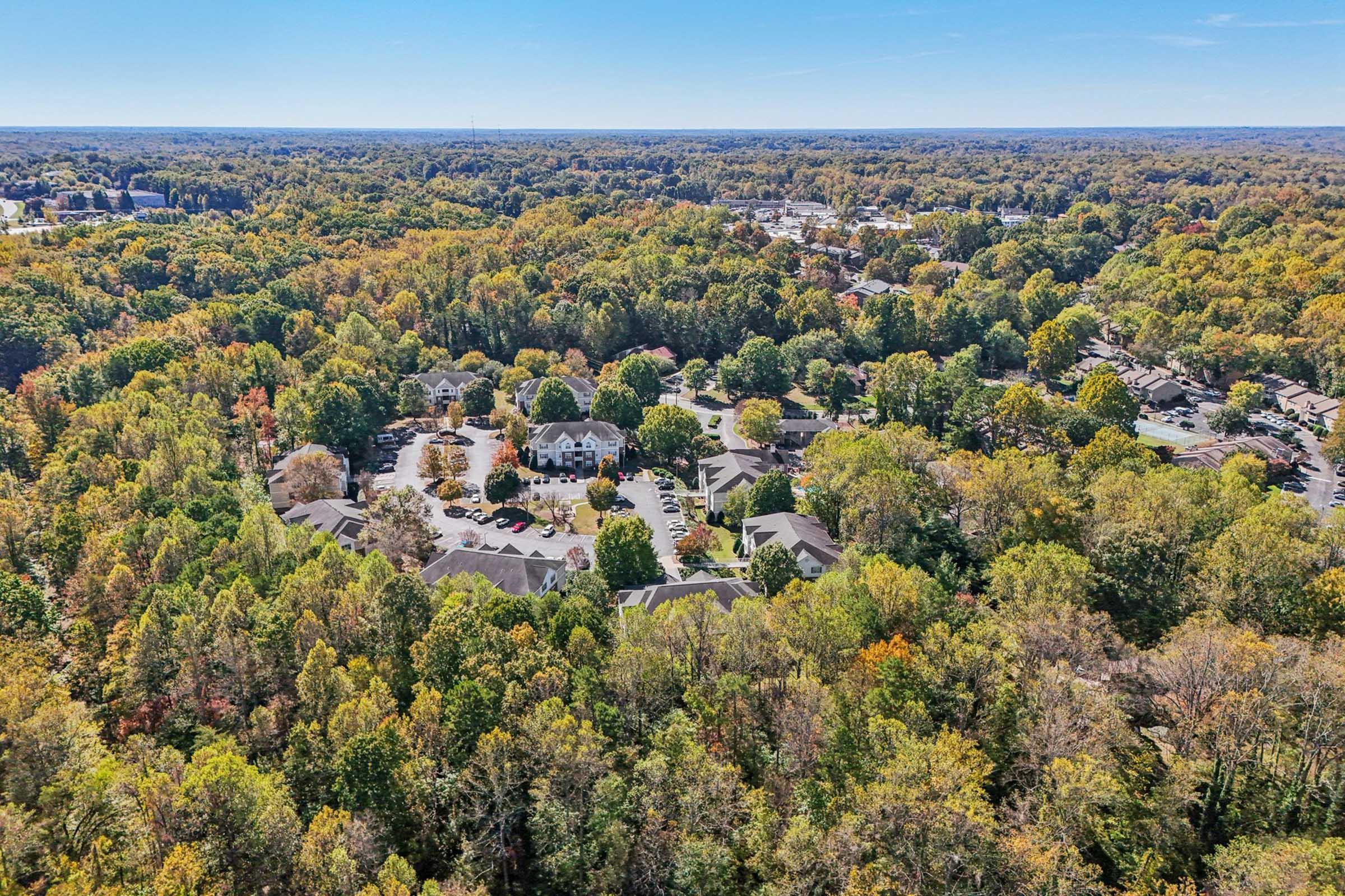 Aerial view of a residential neighborhood surrounded by dense trees in various autumn colors. The layout features multiple buildings in a circular arrangement with parking areas, all set against a backdrop of vibrant fall foliage. Clear blue sky overhead enhances the scenic landscape.