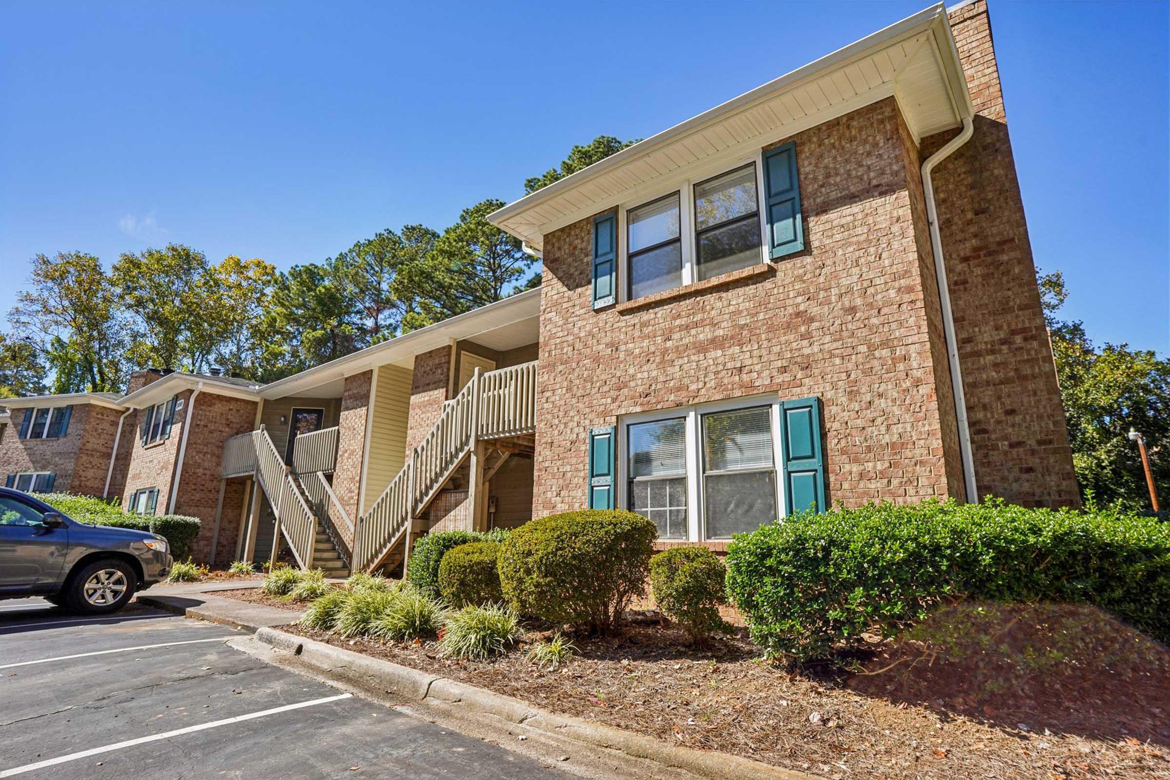 A brick apartment building with white trim and green shutters, featuring a set of stairs leading to upper units. The building is surrounded by manicured shrubs and small plants, with a parking lot in front and trees in the background under a clear blue sky.