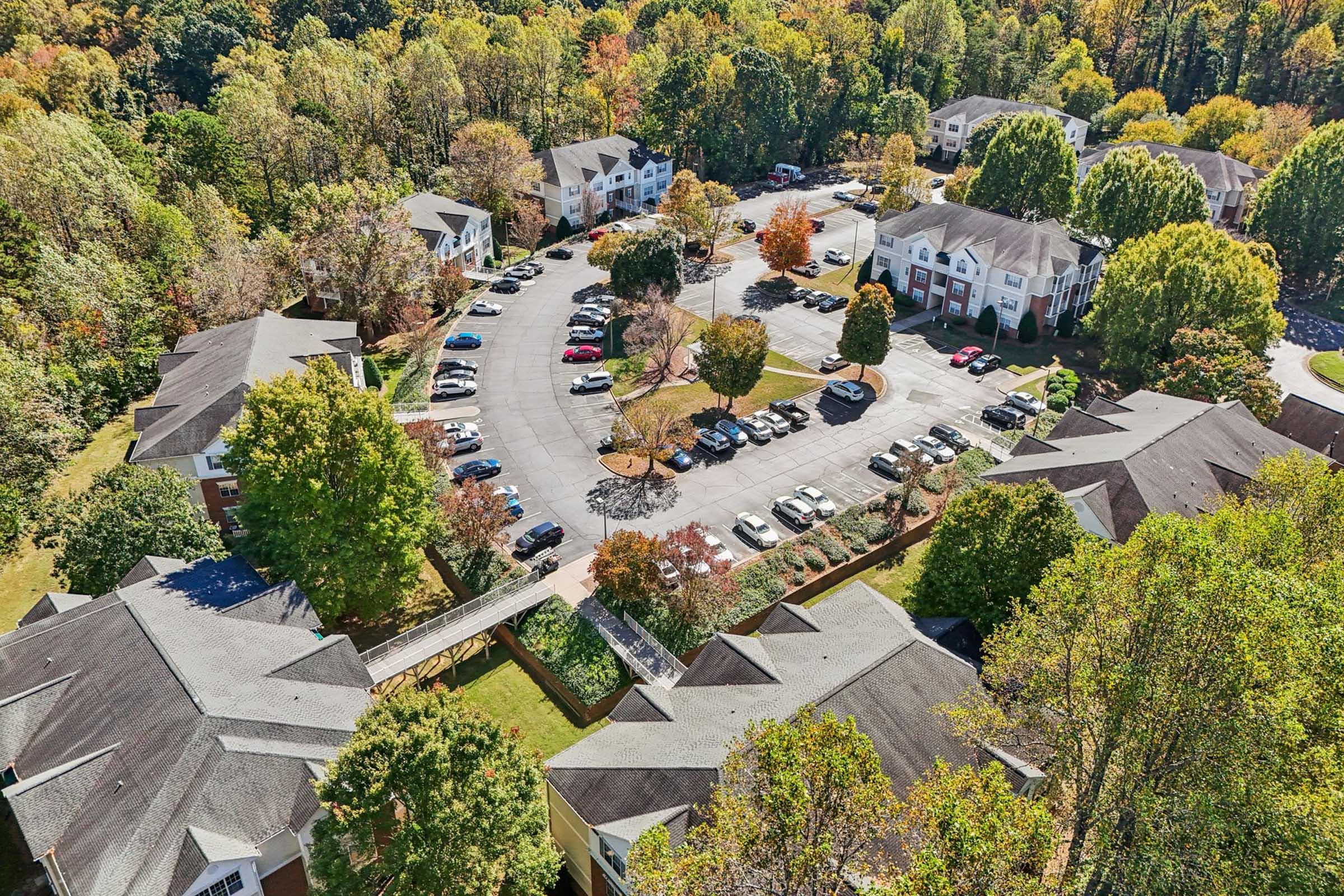 Aerial view of an apartment complex surrounded by lush trees displaying autumn foliage. The scene includes several buildings arranged in a circular layout with a central parking area filled with cars. The surrounding landscape features greenery and colorful fall leaves.